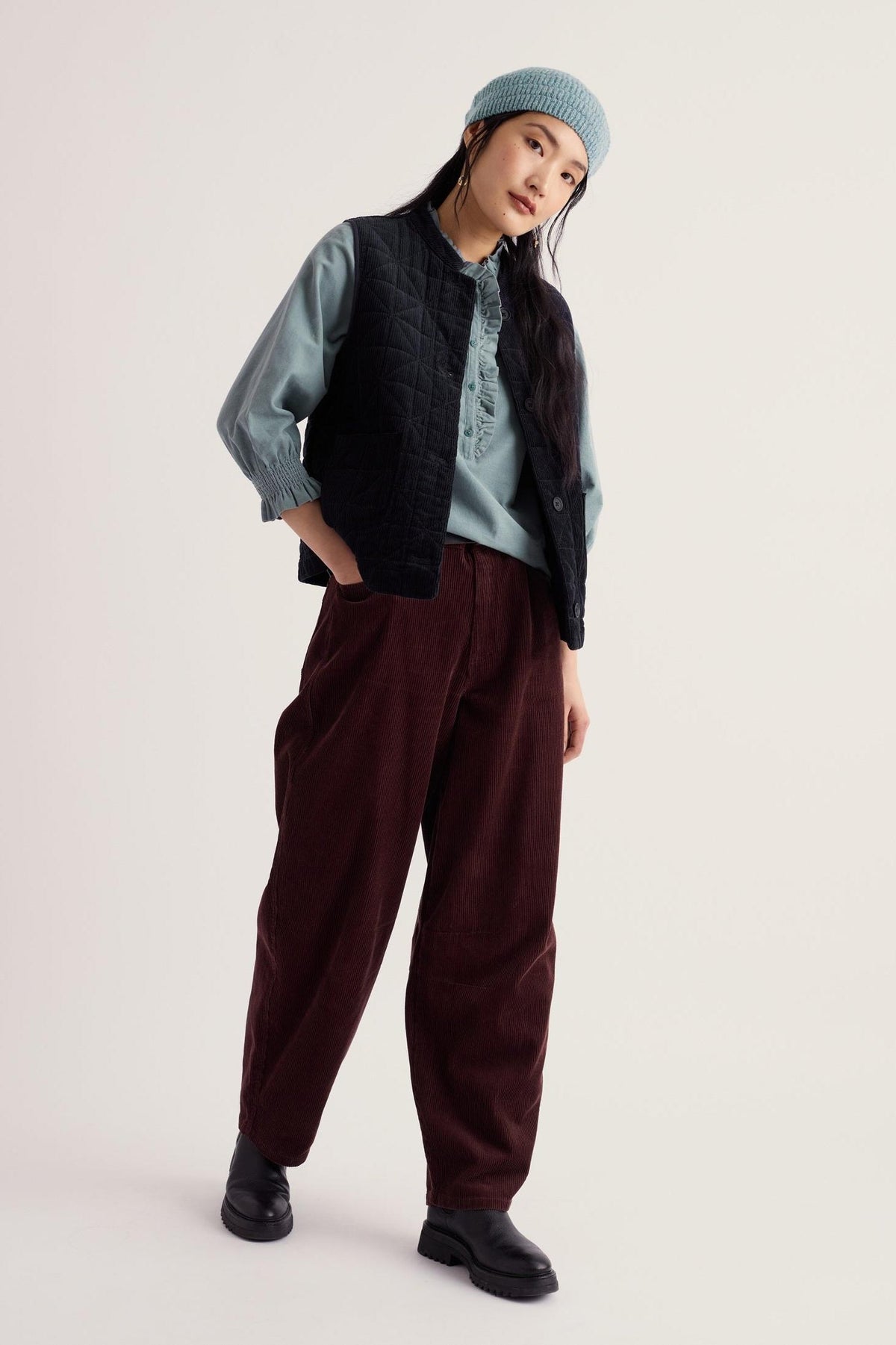 A woman posing in Seasalt Cornwall brown barrel leg cord trousers, a ruffled shirt, quilted vest, and a light blue beanie.