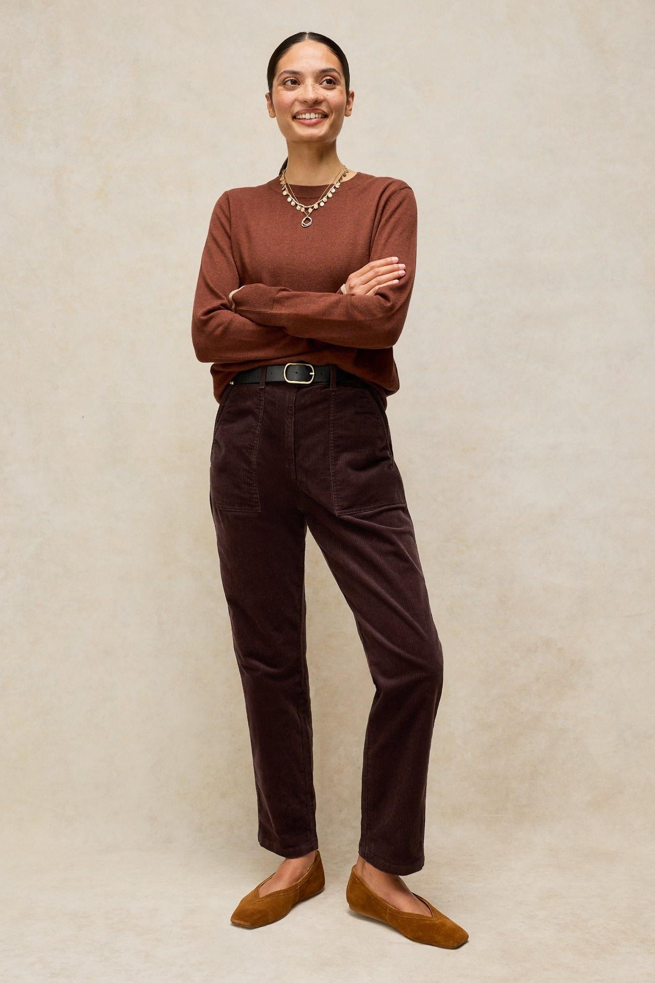 A woman wearing a brown sweater and organic cotton cord trousers standing against a neutral background.
