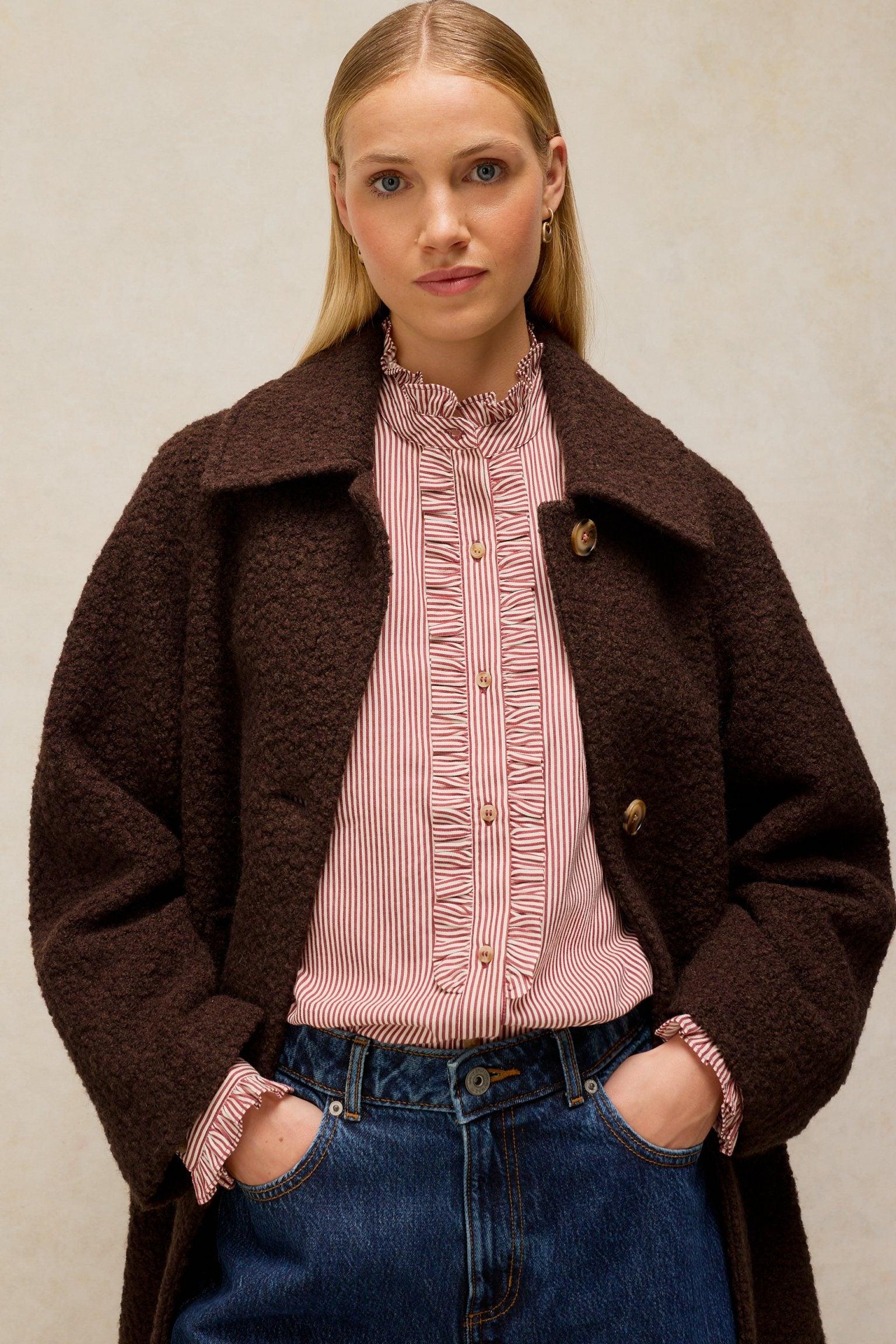 A woman wearing a dark brown textured wool coat over a striped ruffled blouse and blue jeans.