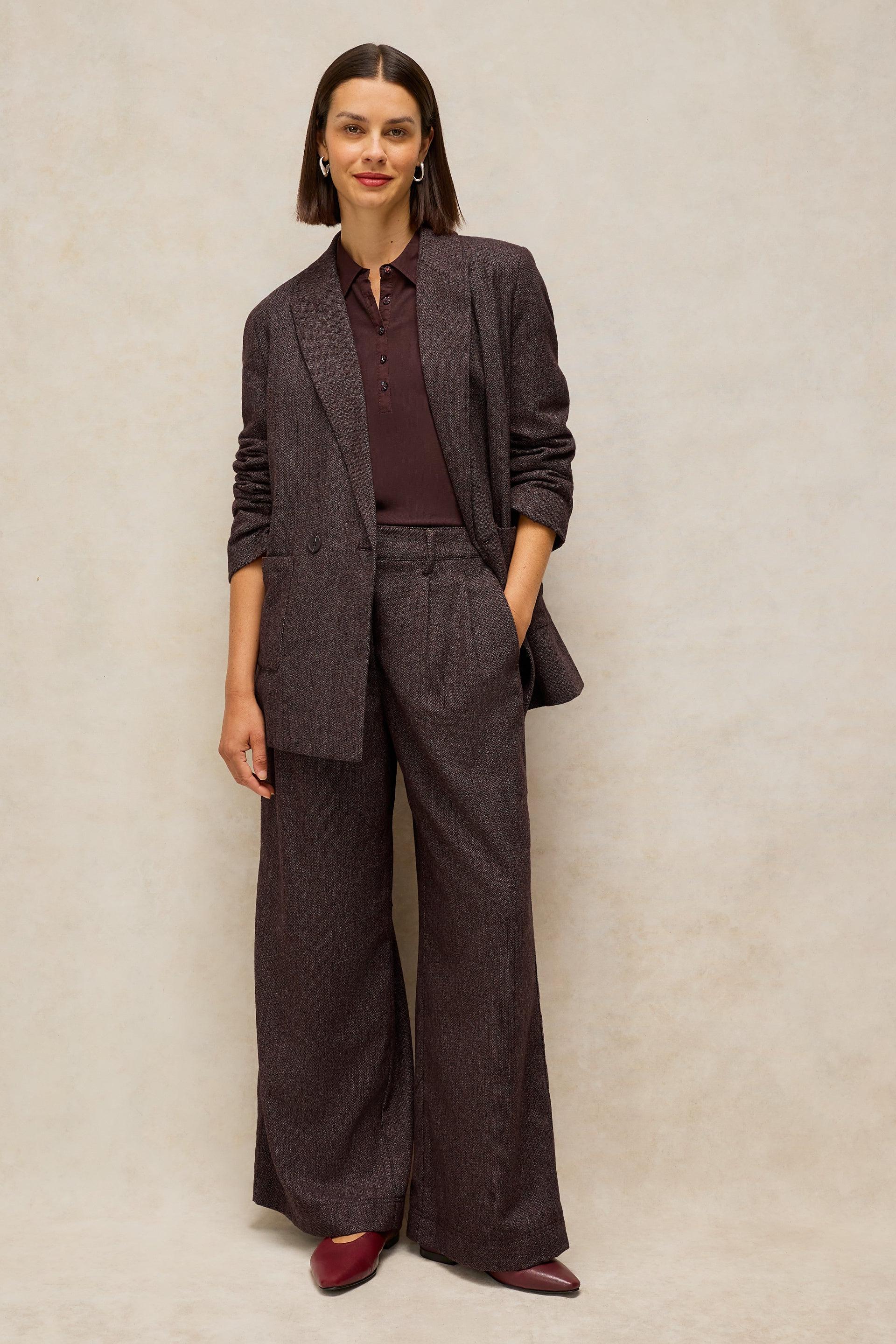 A woman models a dark brown wide-leg blazer and matching trousers set, styled with a dark burgundy collared shirt and burgundy pointed-toe flats, against a textured beige studio backdrop.