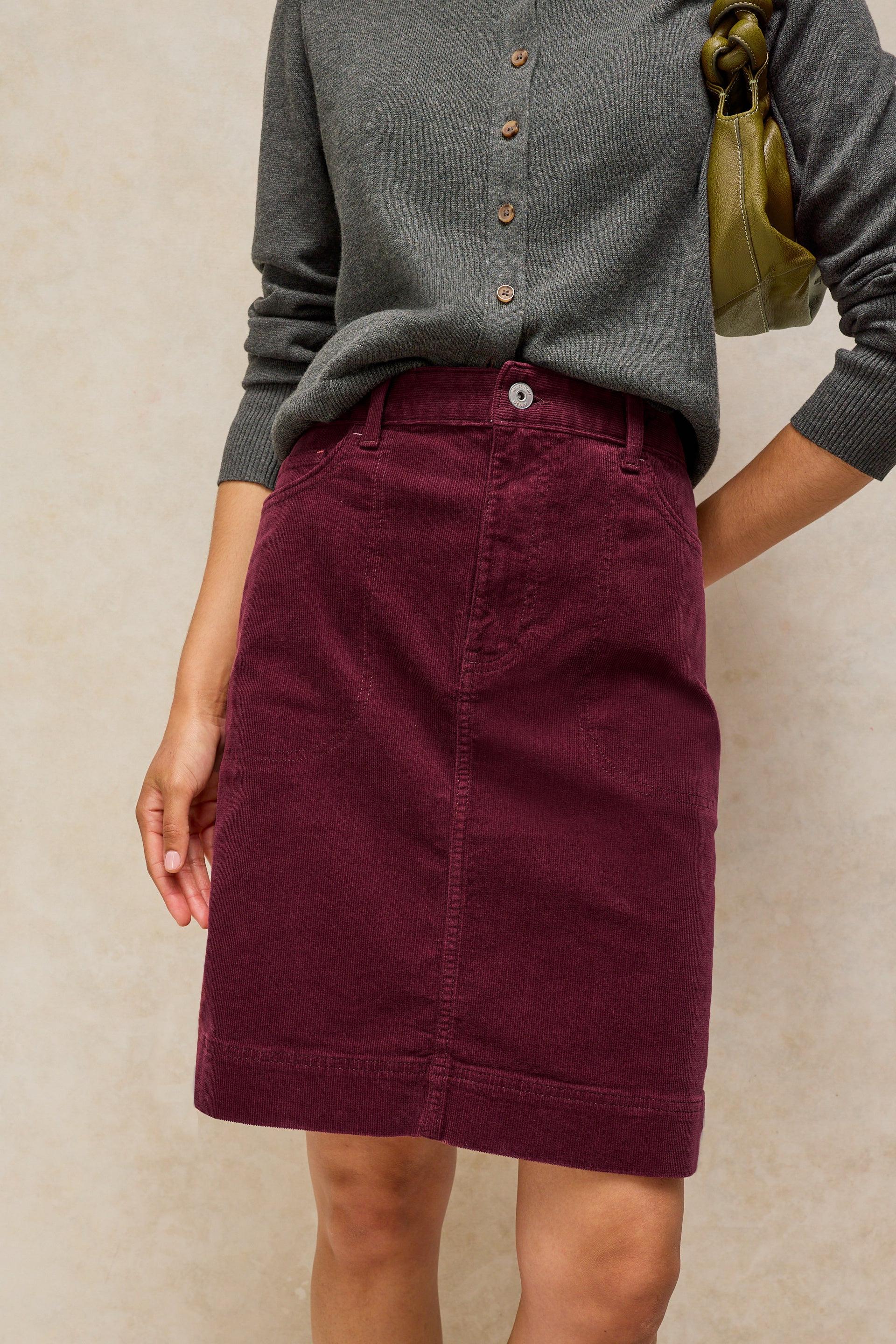 A person wearing a dark burgundy corduroy skirt with a gray button-up cardigan, standing against a textured beige wall.