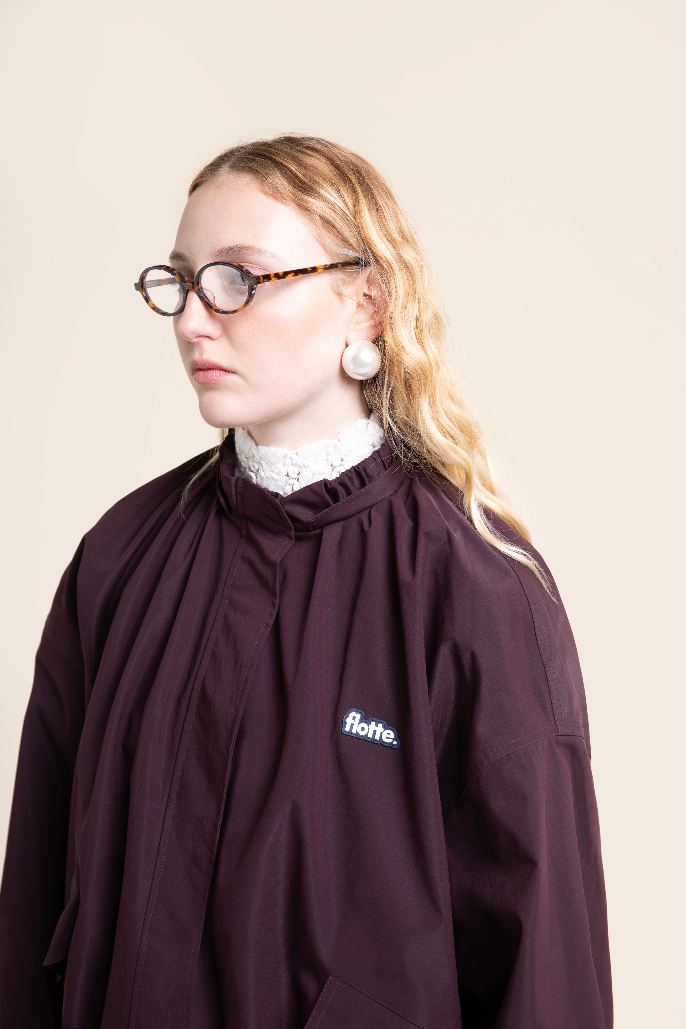 A woman with blonde hair and glasses wearing a burgundy Flotte waterproof coat over a white lace collar.