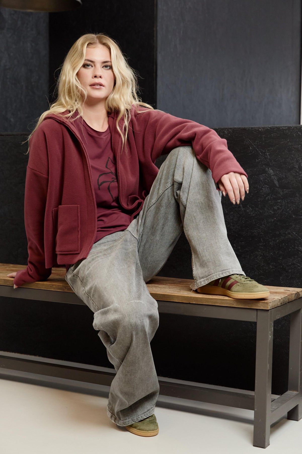 Blonde woman wearing a burgundy Henriette Steffensen fleece cardigan sitting on a wooden bench.