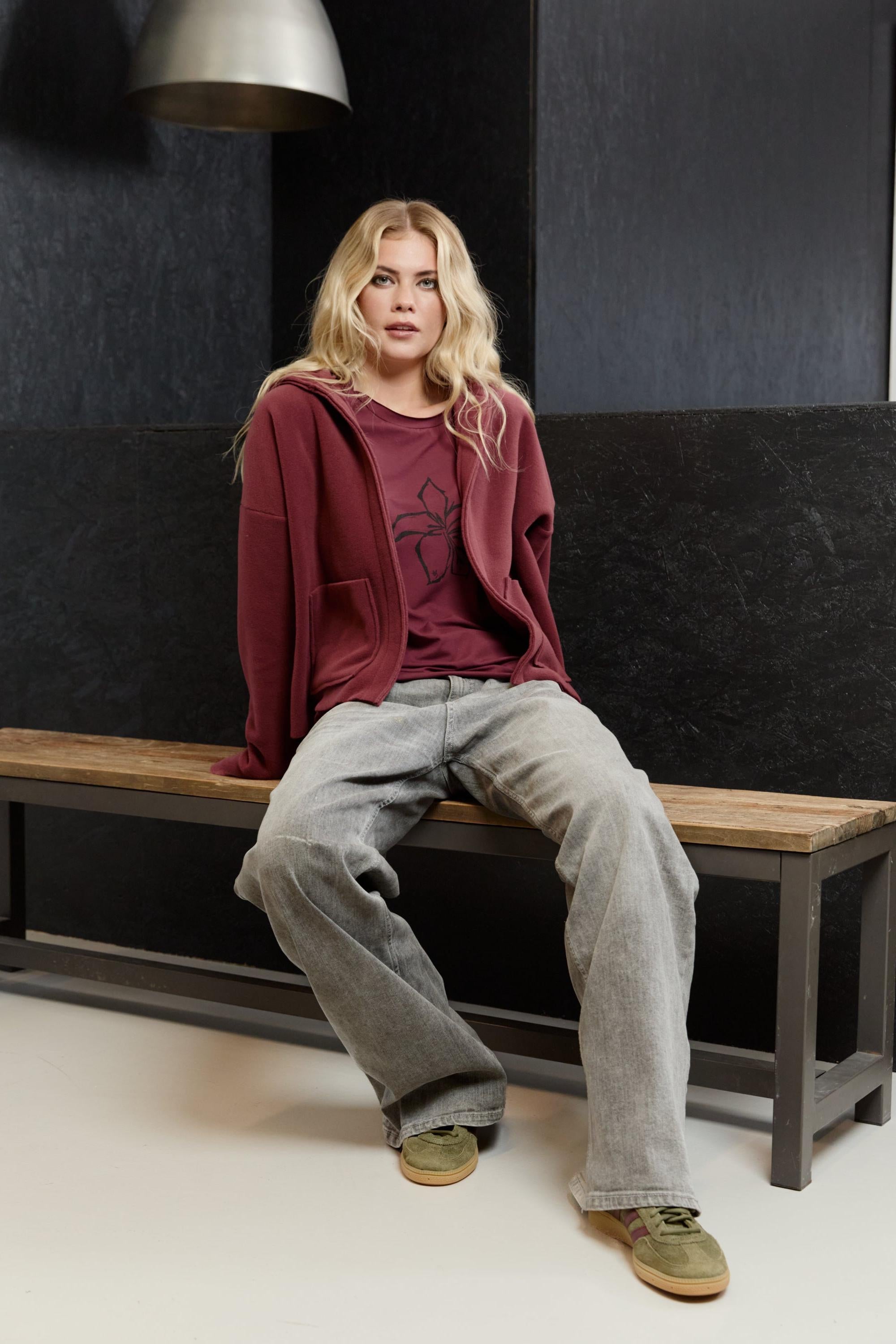 A woman sitting on a wooden bench wearing a burgundy Henriette Steffensen fleece cardigan and casual grey jeans.