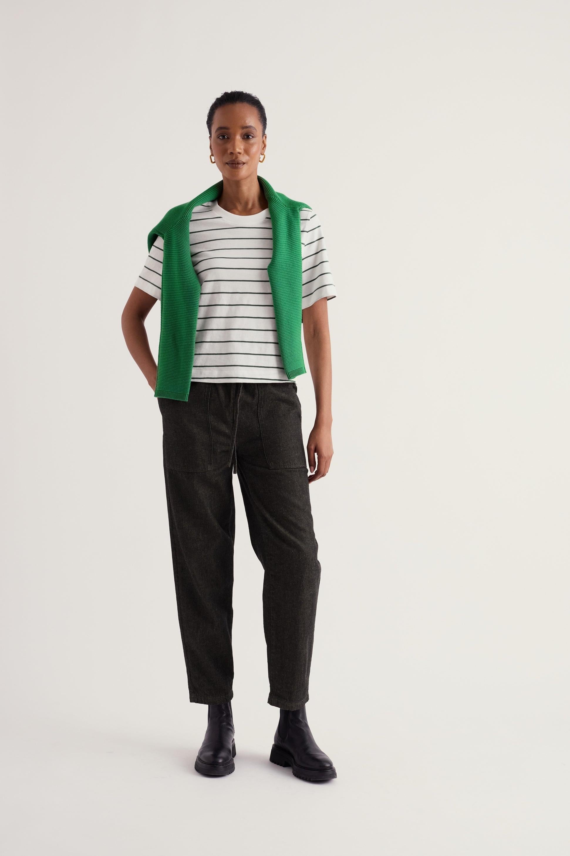 A model wearing black organic cotton trousers and a striped t-shirt with a green sweater draped over her shoulders.