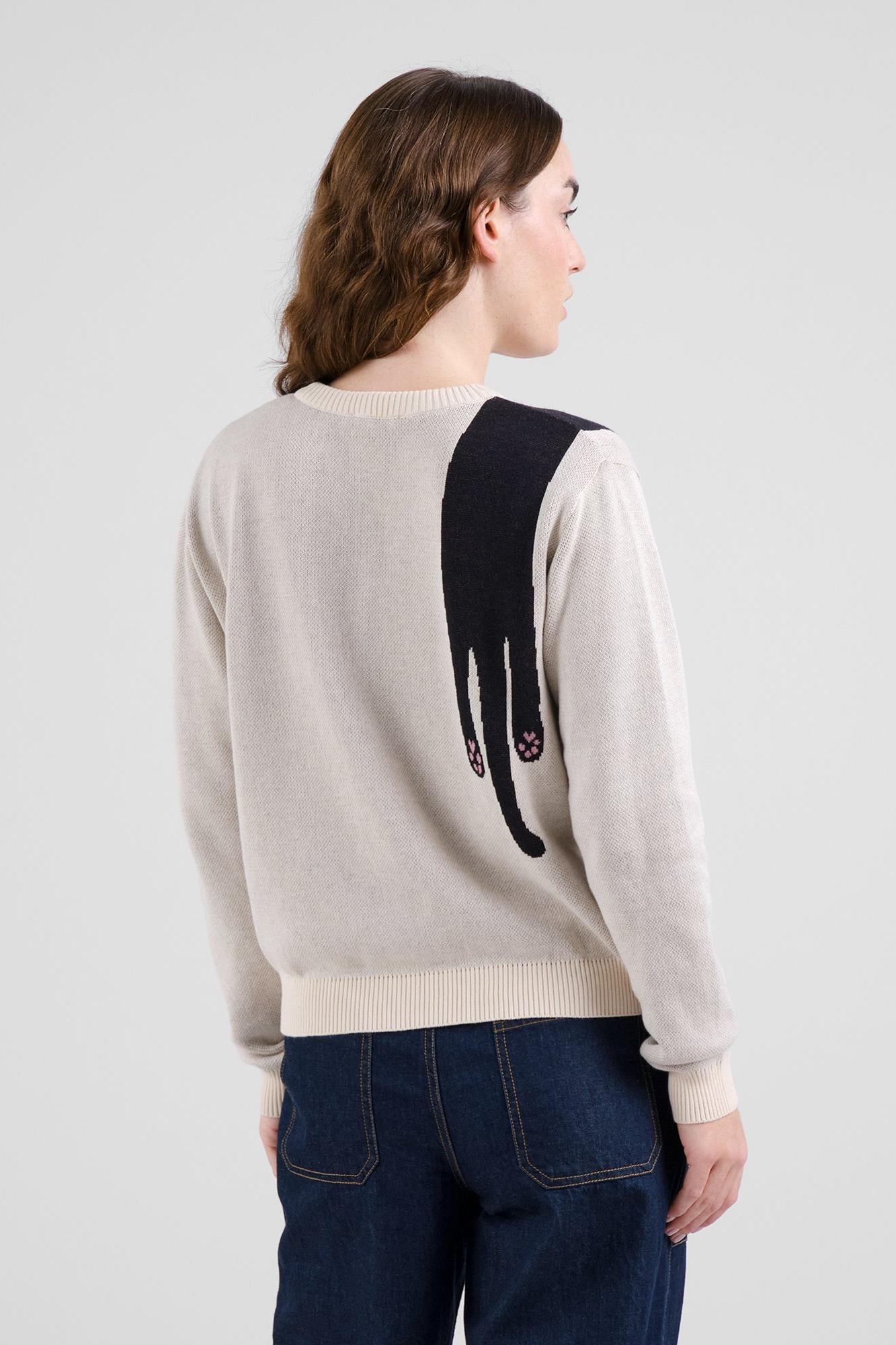 A woman shown from the back wearing a cream-colored knit sweater with a large black cat silhouette graphic on the back, paired with dark blue jeans, against a light gray studio background.