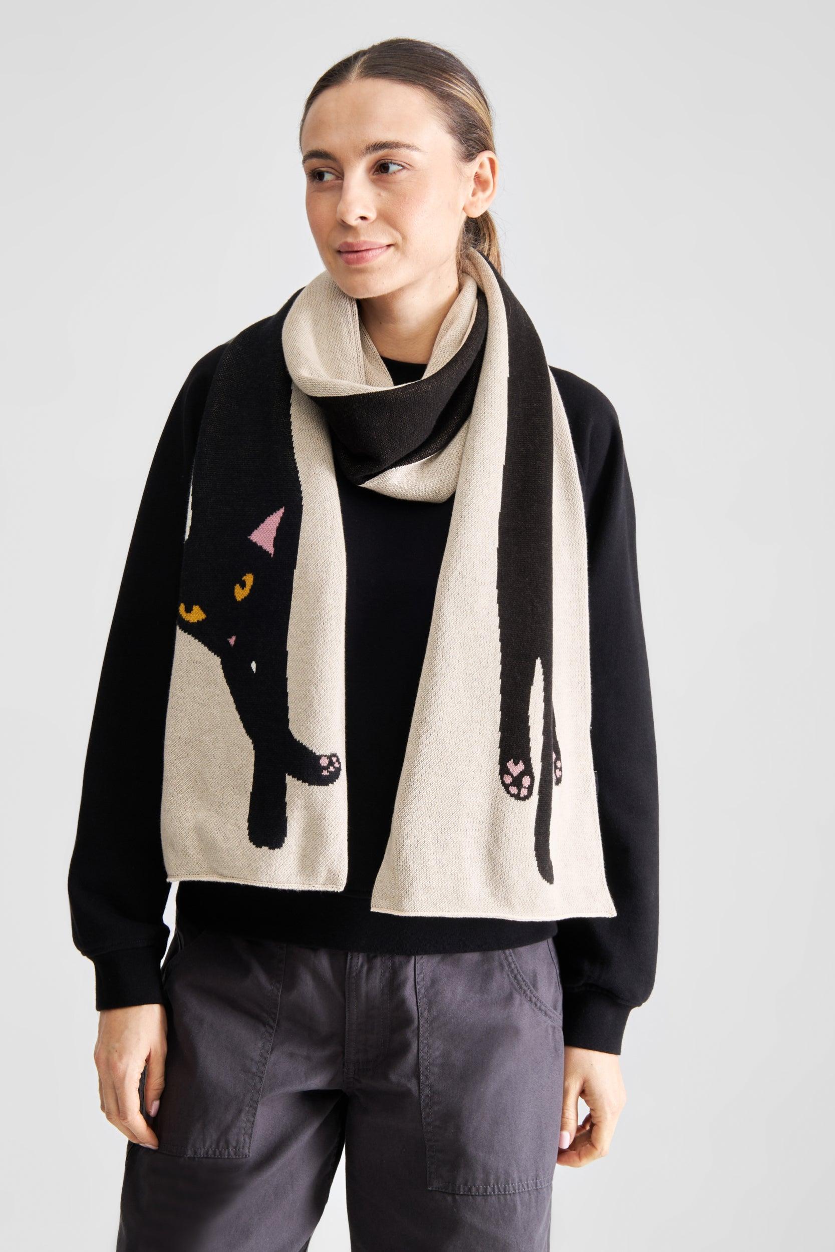 A model wearing a black and cream knit scarf with a black cat graphic, styled over a black sweater and dark gray pants against a light gray studio background.