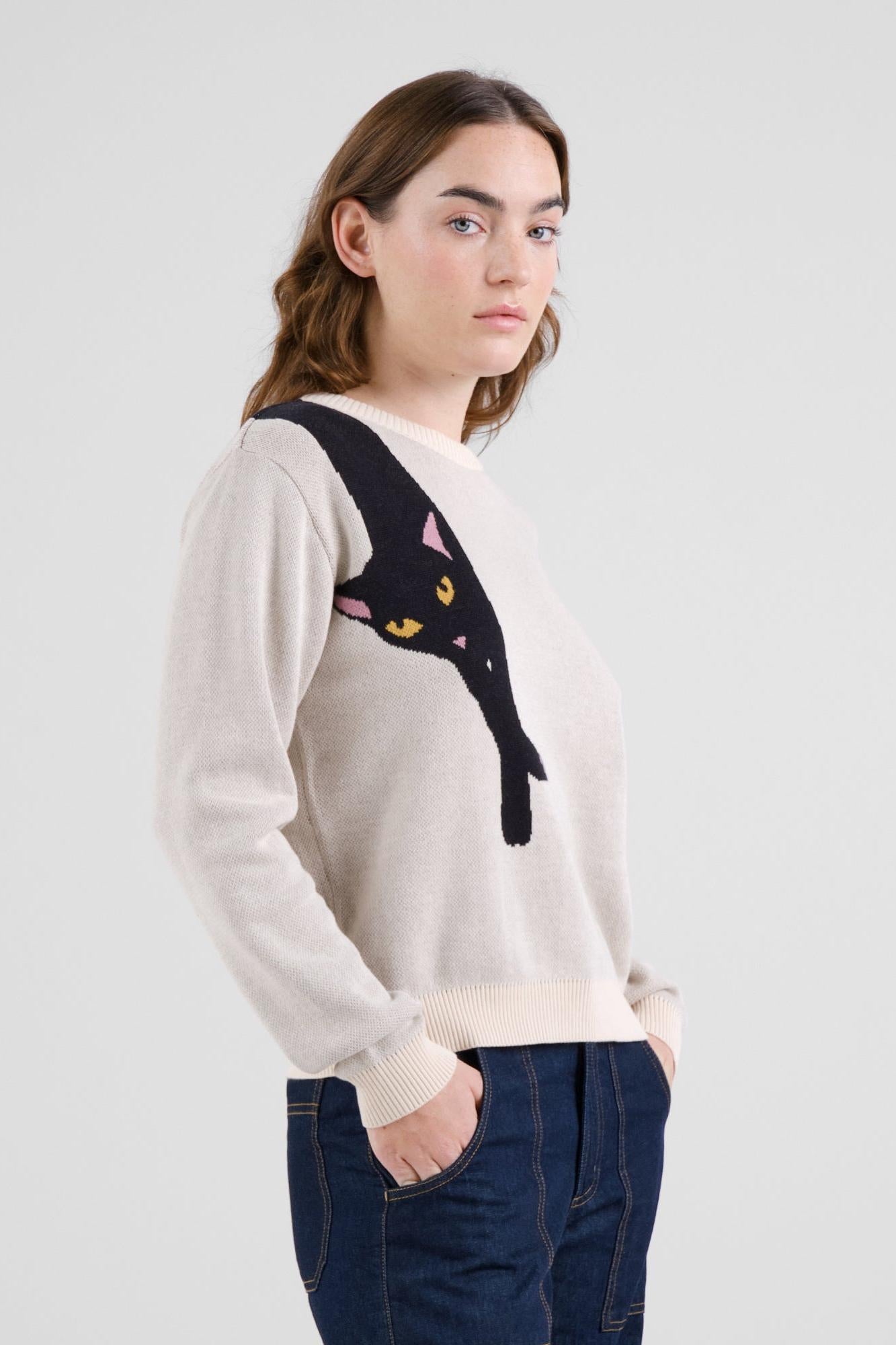A woman wearing a cream-colored knit sweater with a large black cat graphic on the chest, paired with dark blue jeans, photographed against a light gray studio background.
