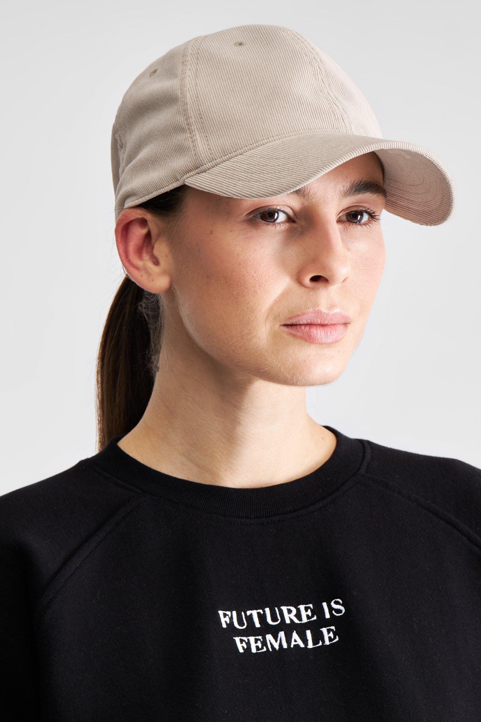 A woman wearing a beige baseball cap and a black crewneck sweatshirt with the text 'FUTURE IS FEMALE' printed in white on the chest, set against a plain light gray background.