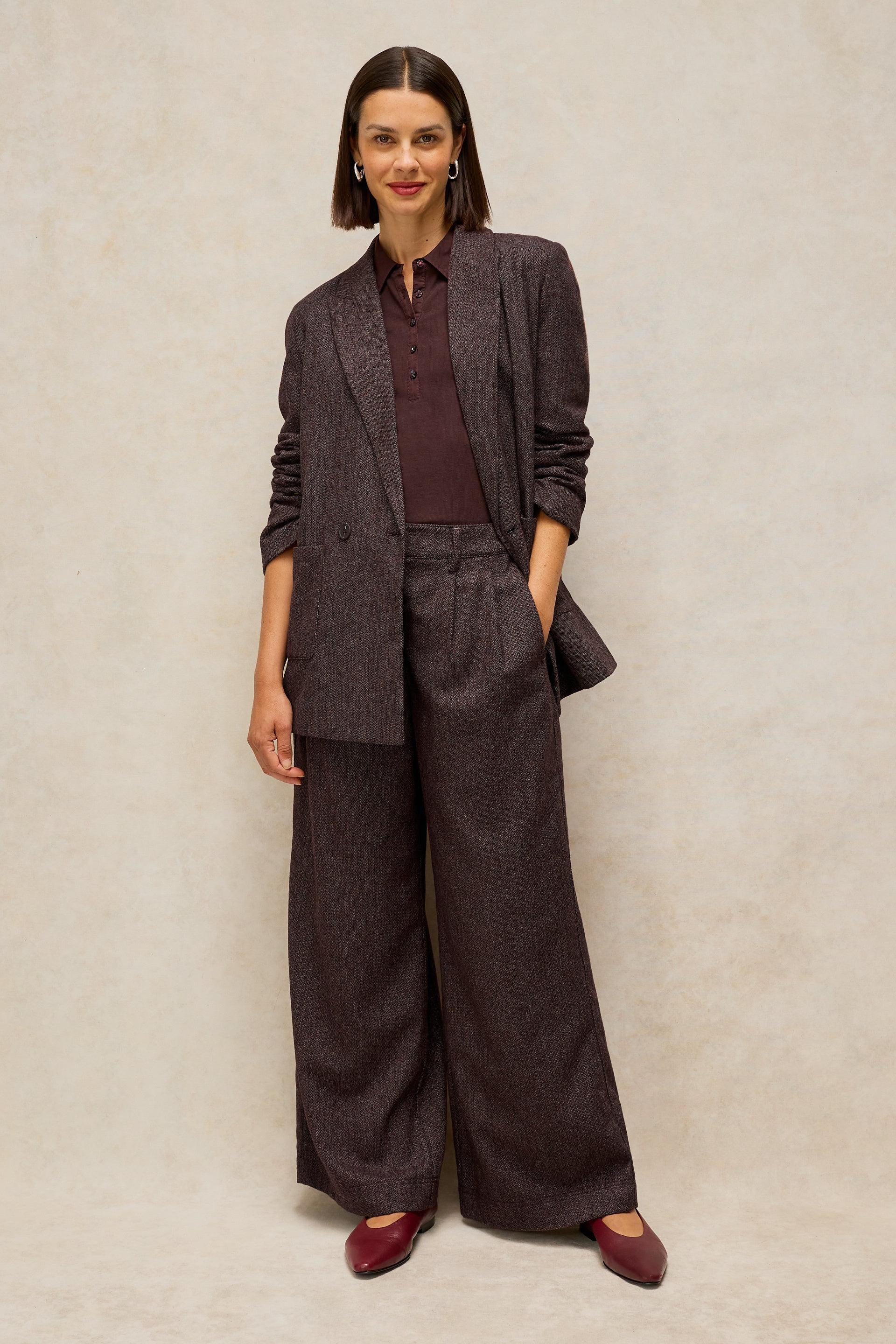 A woman modeling a dark brown two-piece suit with wide-leg trousers and a matching blazer, styled with a burgundy collared shirt and burgundy flats against a textured beige studio backdrop.