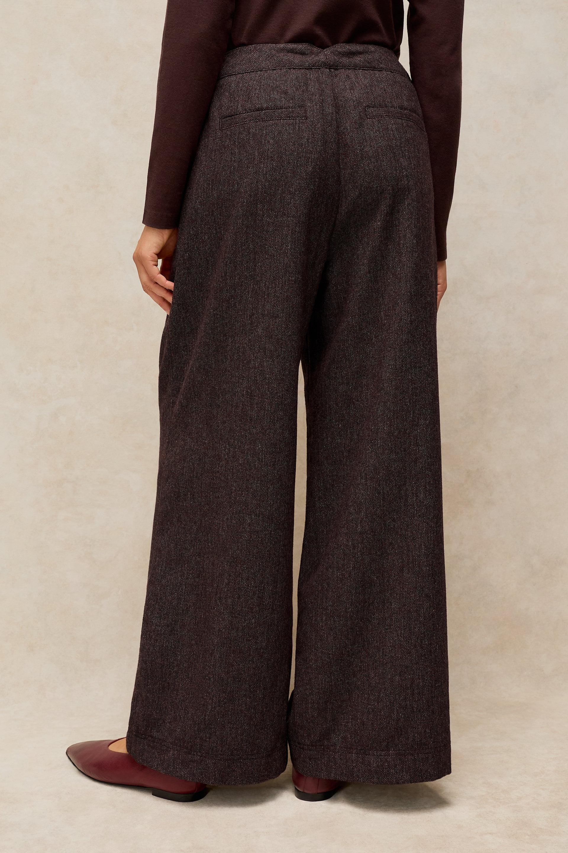 A person shown from the back wearing dark brown wide-leg trousers, a dark brown top, and burgundy flats against a textured beige studio backdrop.