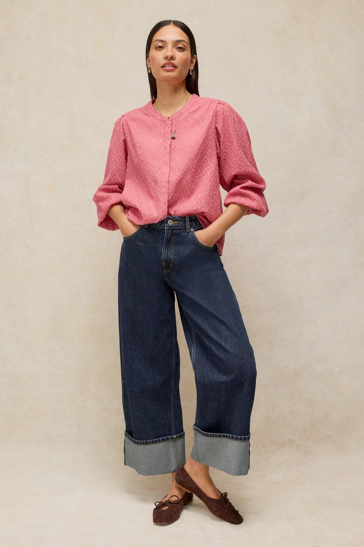 A woman posing in dark denim jeans with wide cuffs and a textured pink blouse against a neutral background.
