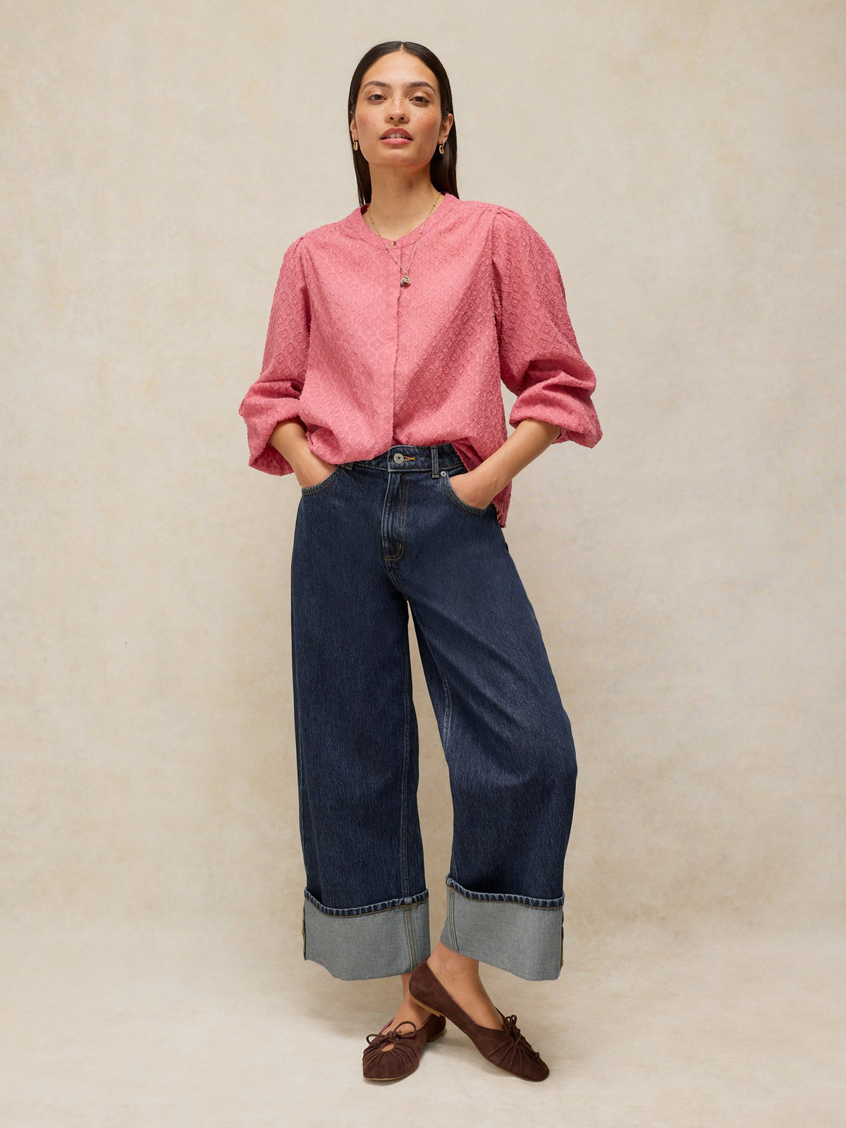A woman posing in dark denim jeans with wide cuffs and a textured pink blouse against a neutral background.