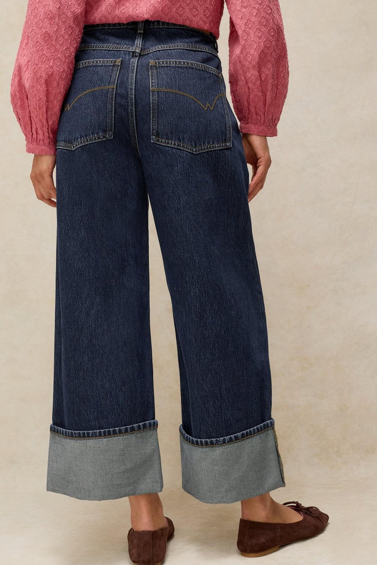 Rear view of a woman wearing dark denim jeans with wide cuffed hems paired with a pink textured blouse.