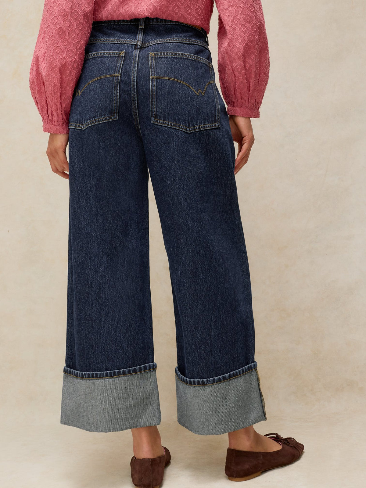 Rear view of a woman wearing dark denim jeans with wide cuffed hems paired with a pink textured blouse.