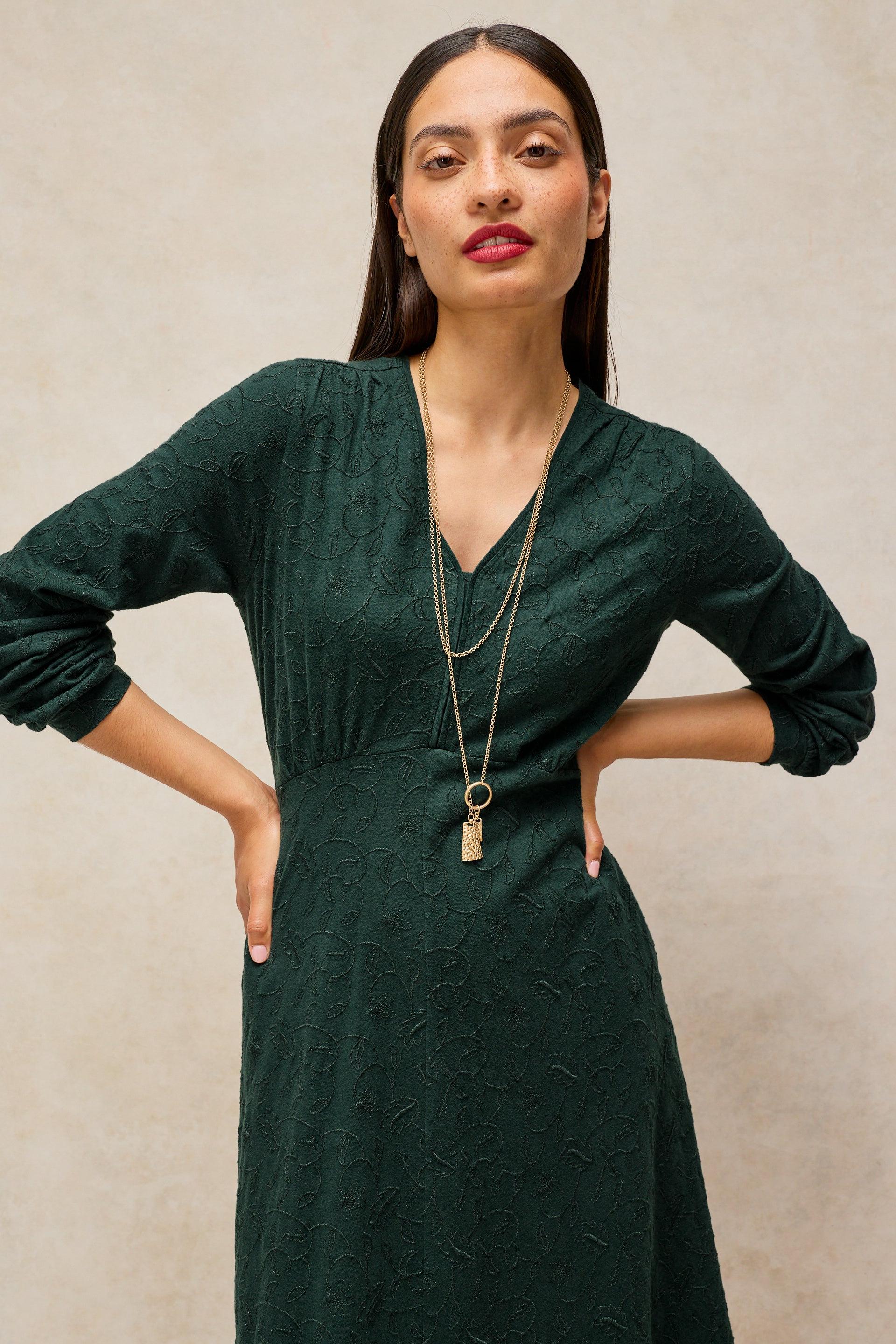 A woman modeling a dark forest green textured long-sleeve dress with a V-neckline, paired with a double-layered gold pendant necklace, set against a neutral beige textured wall.