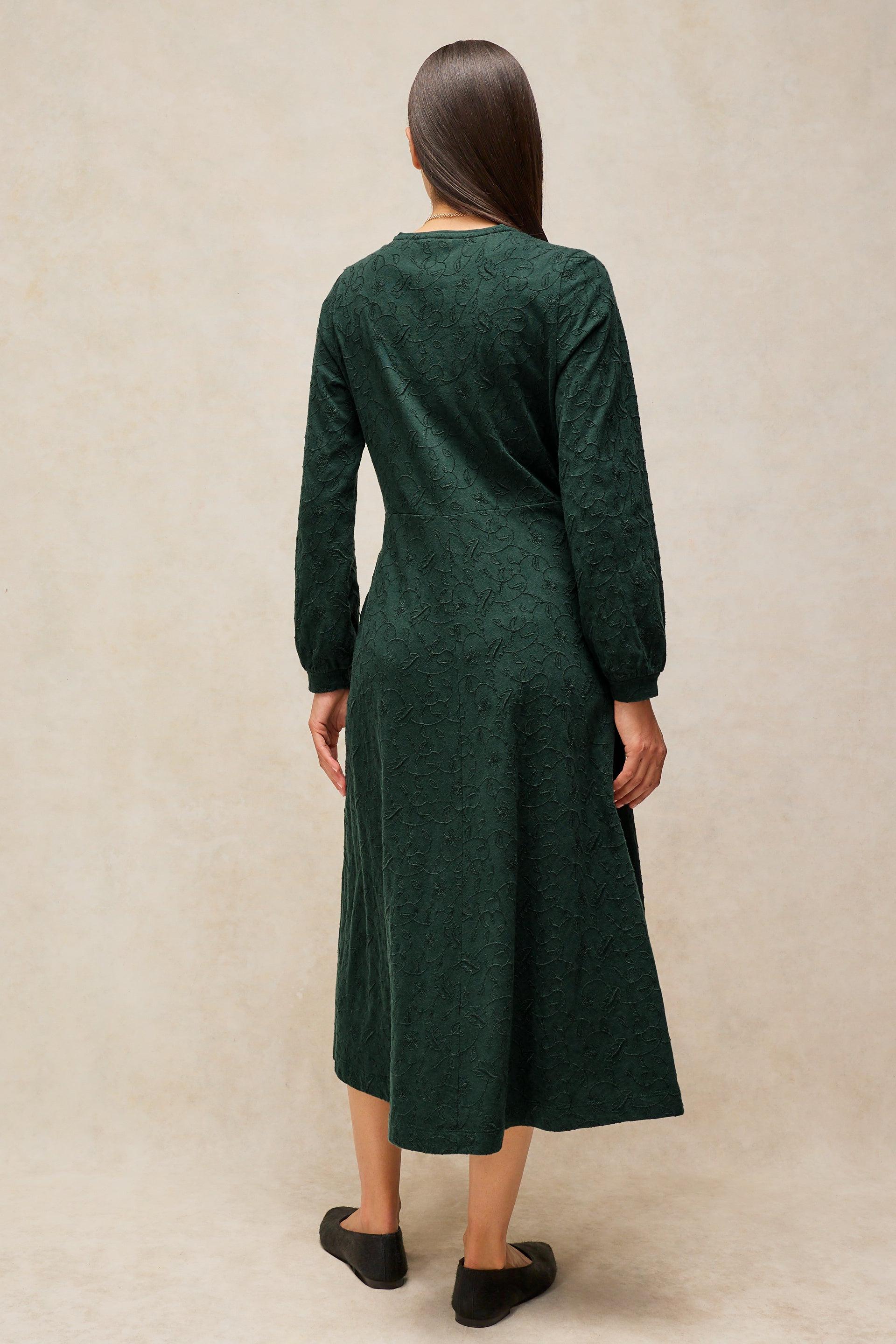 A woman viewed from the back wearing a dark green long-sleeve midi dress with a textured knit pattern, standing against a neutral beige studio backdrop.
