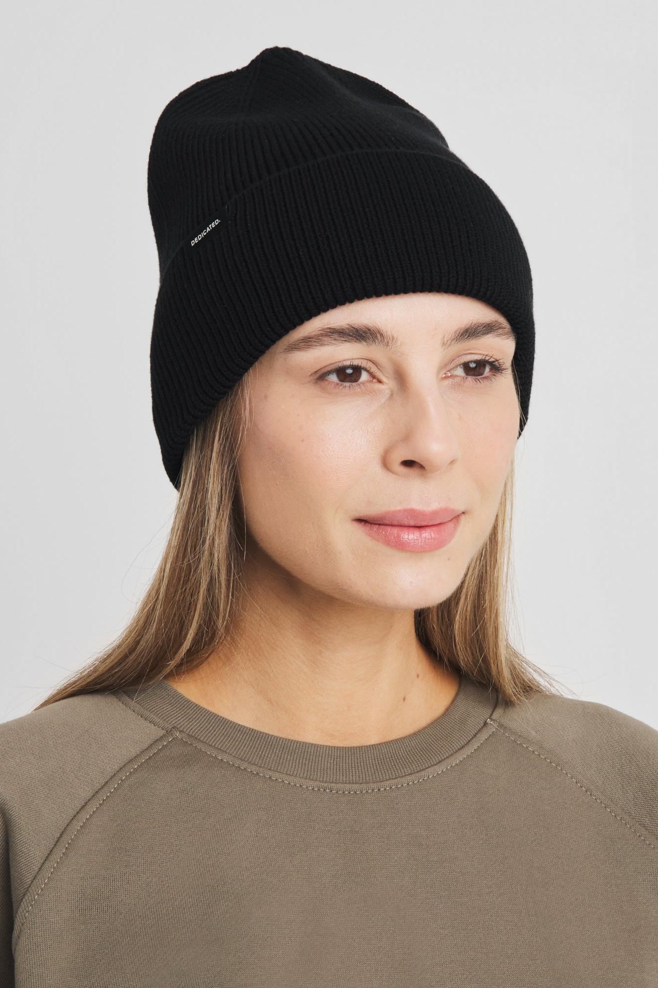 A woman wearing a black ribbed knit beanie and an olive green sweatshirt, photographed against a neutral light gray background.