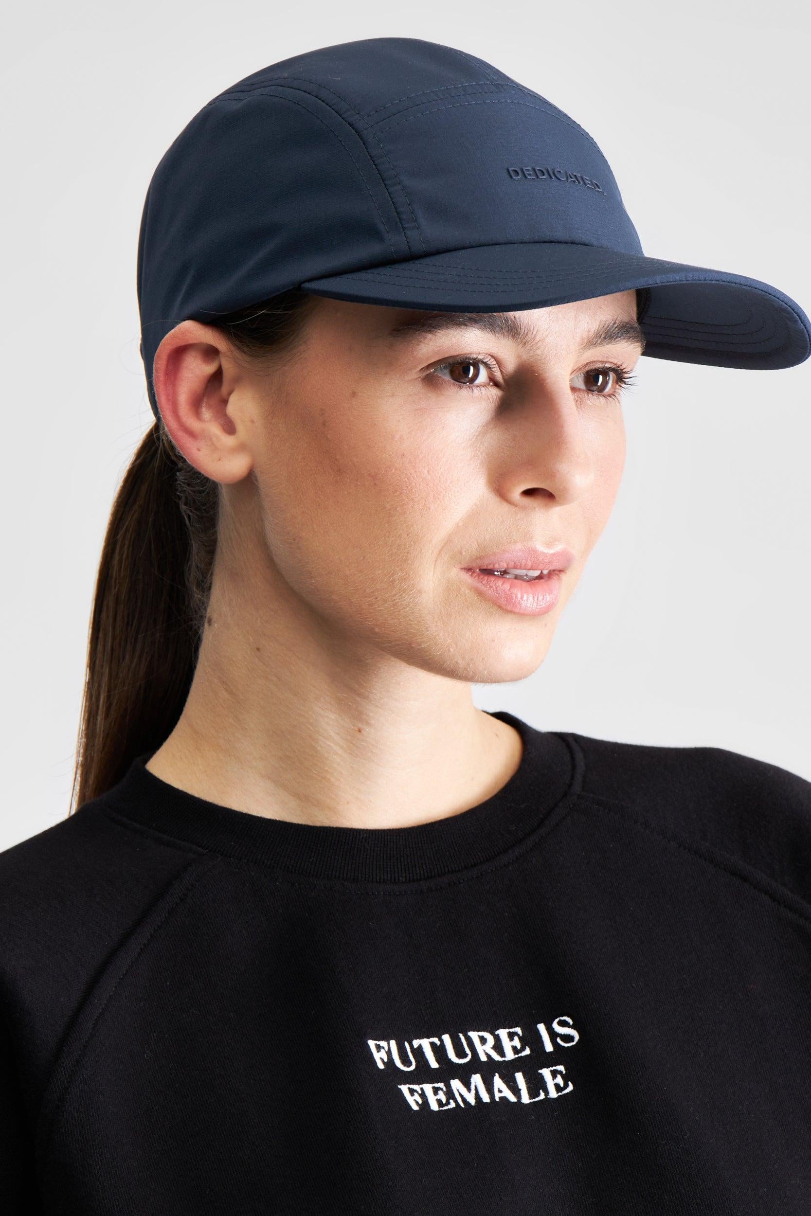 A woman wearing a navy blue five-panel cap with 'DEDICATED' embroidered on the side and a black crewneck sweatshirt with 'FUTURE IS FEMALE' printed in white on the chest, set against a plain light gray background.