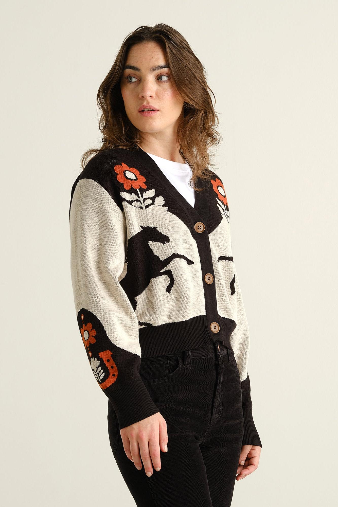 A woman modeling a black and cream knit cardigan with orange floral and black graphic motifs, paired with black pants against a plain light background.