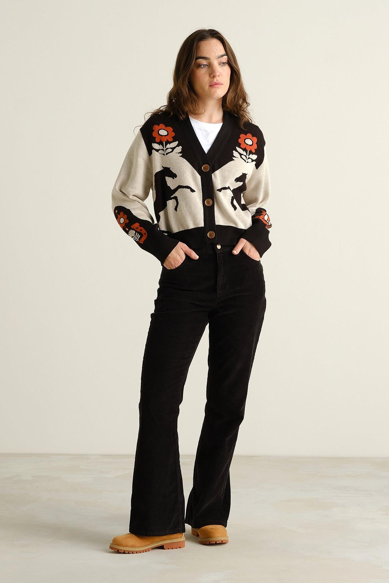 A woman stands in a studio wearing a cream and black knit cardigan with orange and red floral motifs, black flared pants, and tan boots against a plain light background.