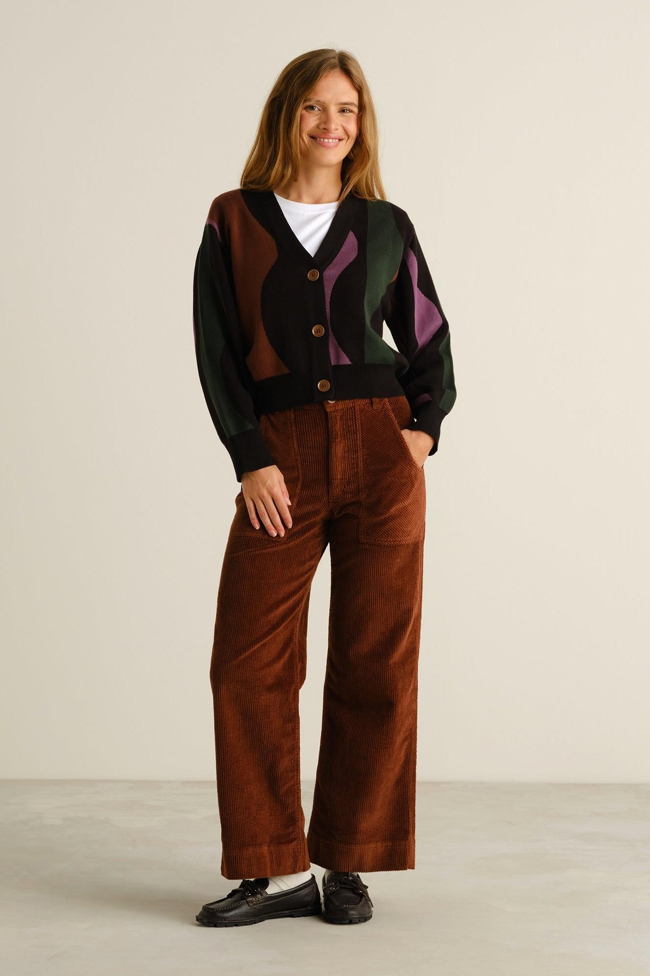 A woman stands in a studio wearing a cropped cardigan with abstract color-blocked panels in black, brown, green, and purple over a white top, paired with wide-leg brown corduroy pants and black shoes.