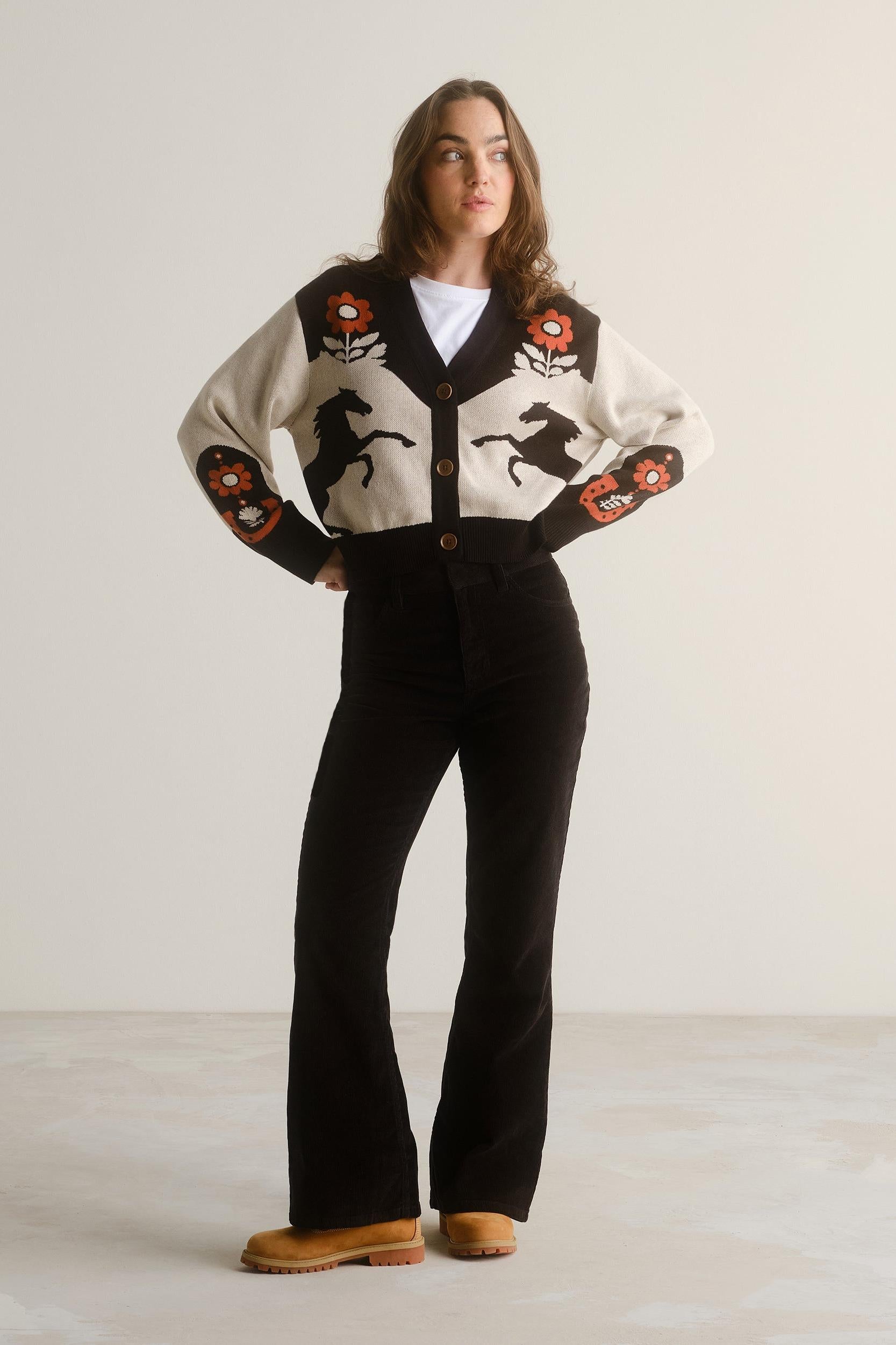A woman stands in a studio wearing a cream knit cardigan with black panels and embroidered red, orange, and black floral/horse motifs, paired with black pants and tan boots.