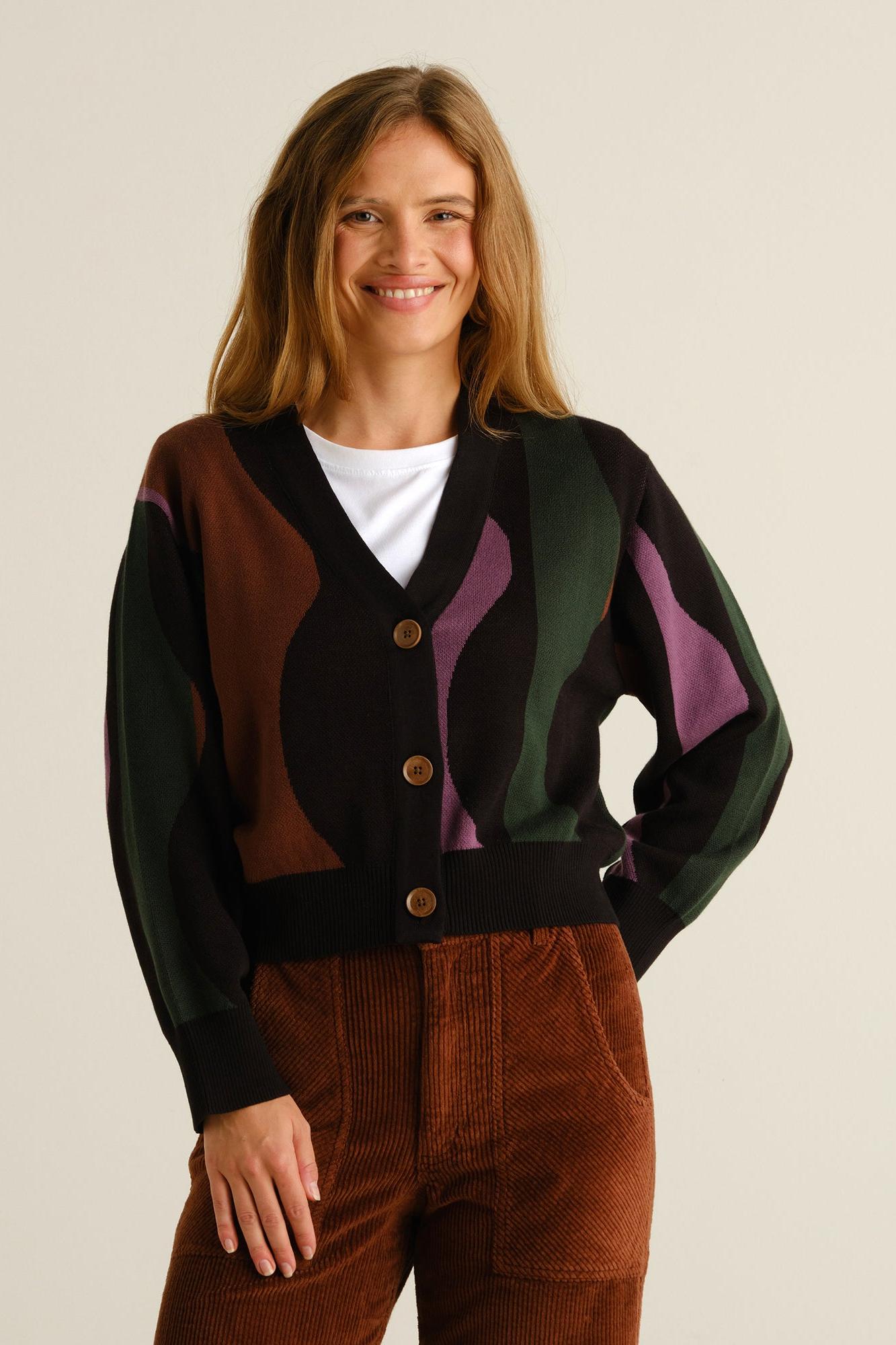 A woman modeling a black cardigan with abstract wavy color-blocked panels in brown, dark green, and purple, worn over a white t-shirt and brown corduroy pants against a light beige background.