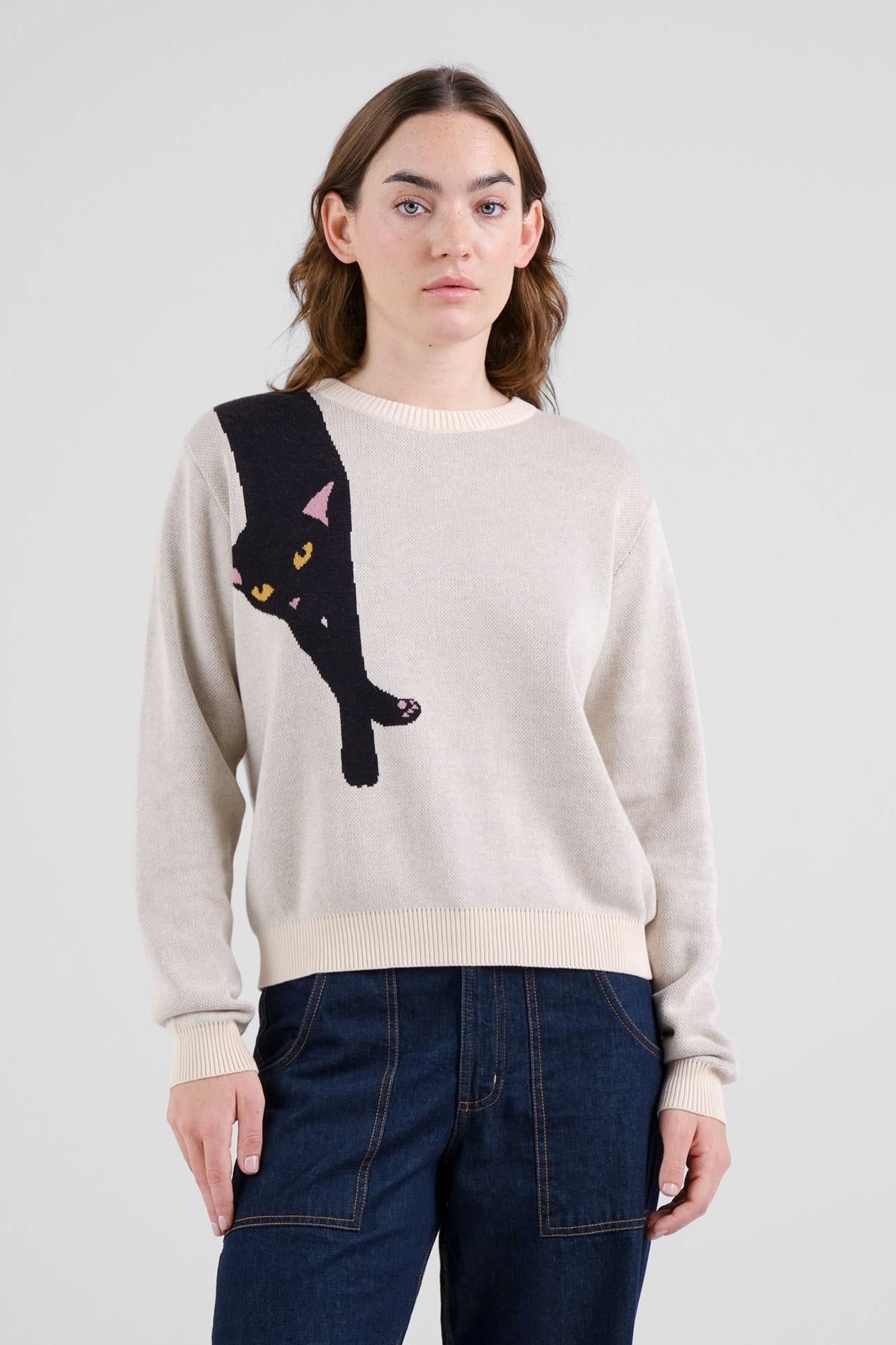 A woman wearing a light beige crewneck sweater with a large black cat graphic on the front, paired with dark blue jeans, against a plain light gray studio background.