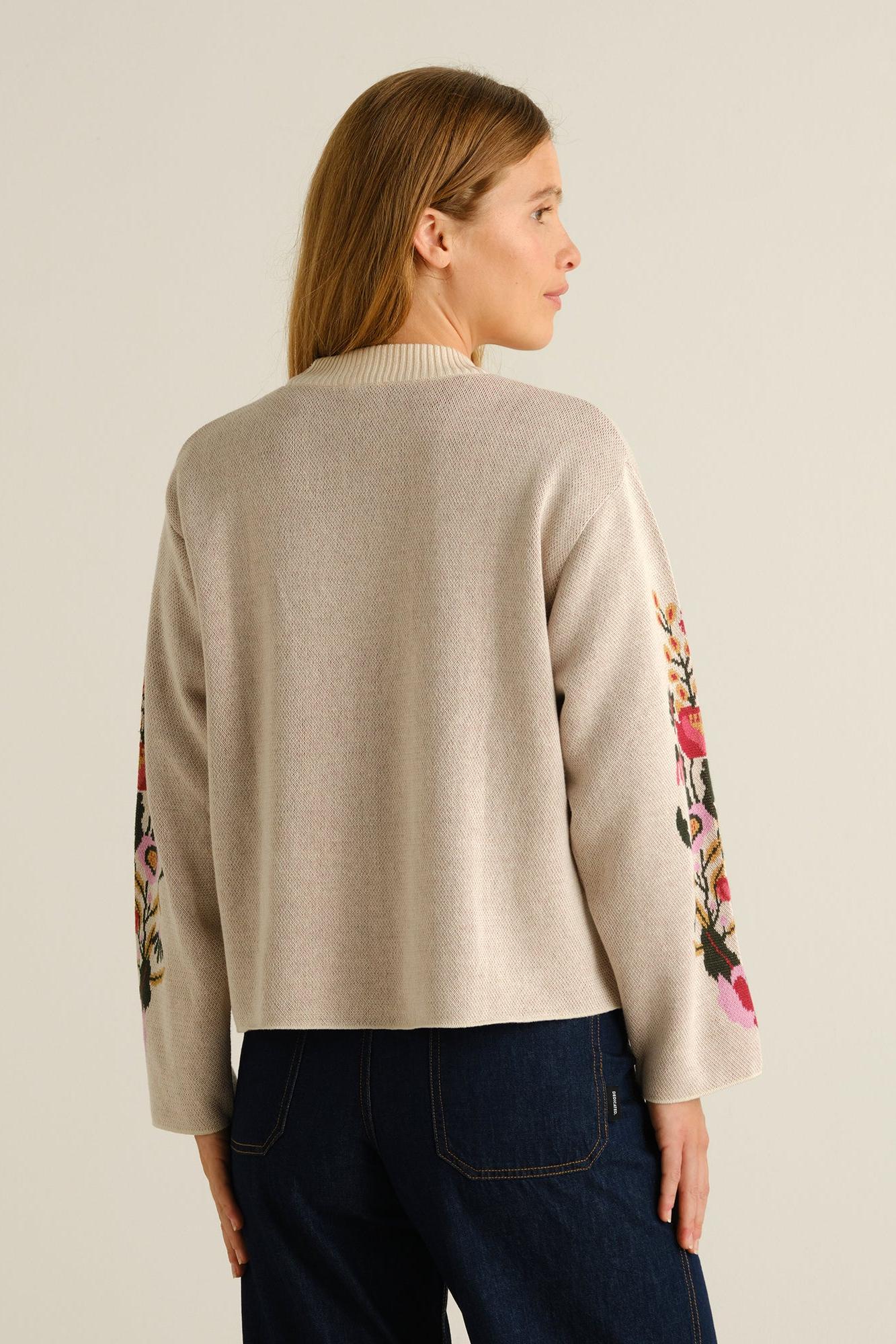 A woman shown from the back wearing a light beige knit sweater with colorful floral embroidery on the sleeves, paired with dark blue jeans, against a plain light beige background.