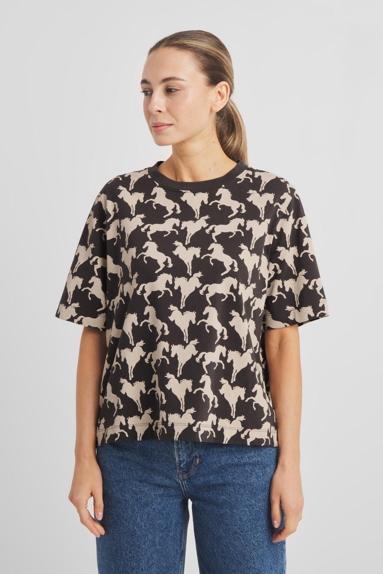 A woman wearing a short-sleeve crewneck t-shirt with an allover black and beige horse pattern, paired with blue jeans, photographed against a neutral light gray background.