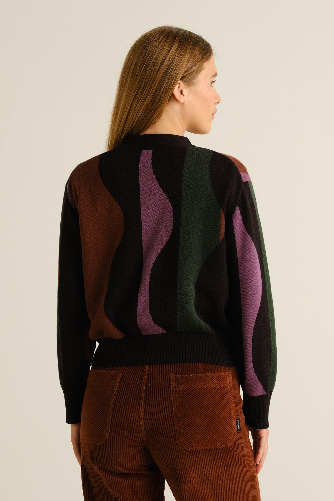 A woman shown from the back wearing a black knit sweater with large abstract wavy panels in brown, green, and purple, paired with brown corduroy pants, against a plain light beige studio background.