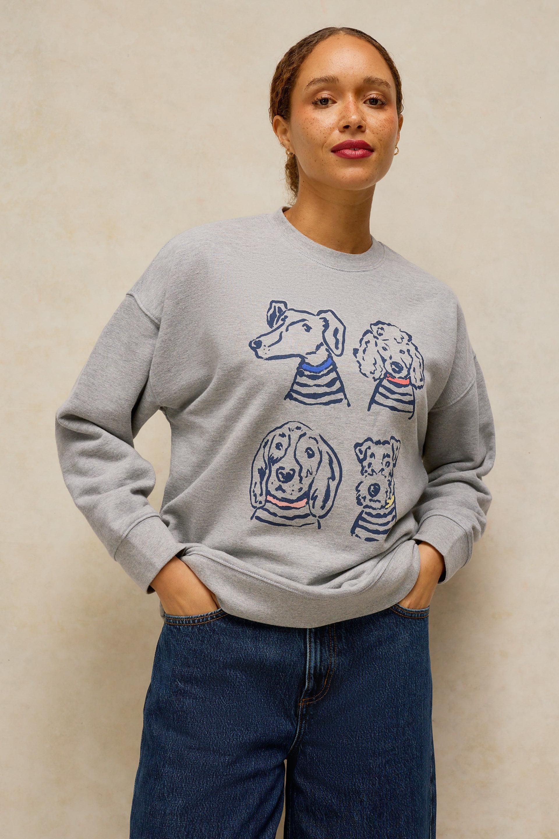 A woman wearing a light gray crewneck sweatshirt with a graphic print of four dogs wearing striped shirts, paired with dark blue jeans, standing against a textured beige wall.