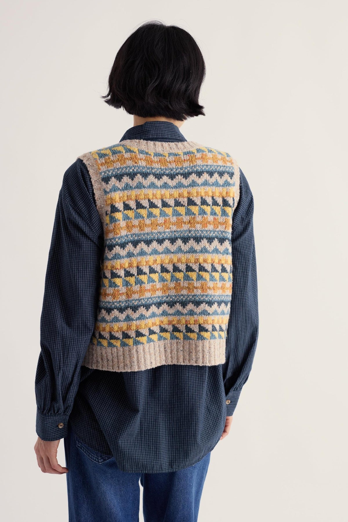 Woman seen from behind wearing a colourful Seasalt Cornwall Fair Isle knit vest over a navy checkered shirt.