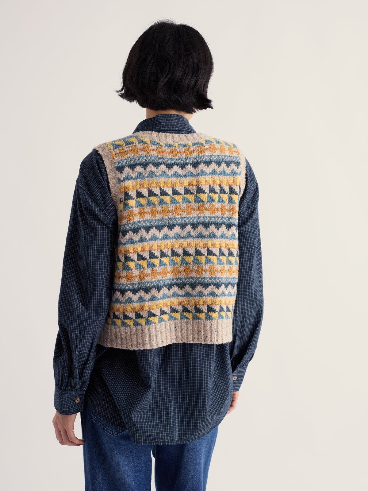 Woman seen from behind wearing a colourful Seasalt Cornwall Fair Isle knit vest over a navy checkered shirt.