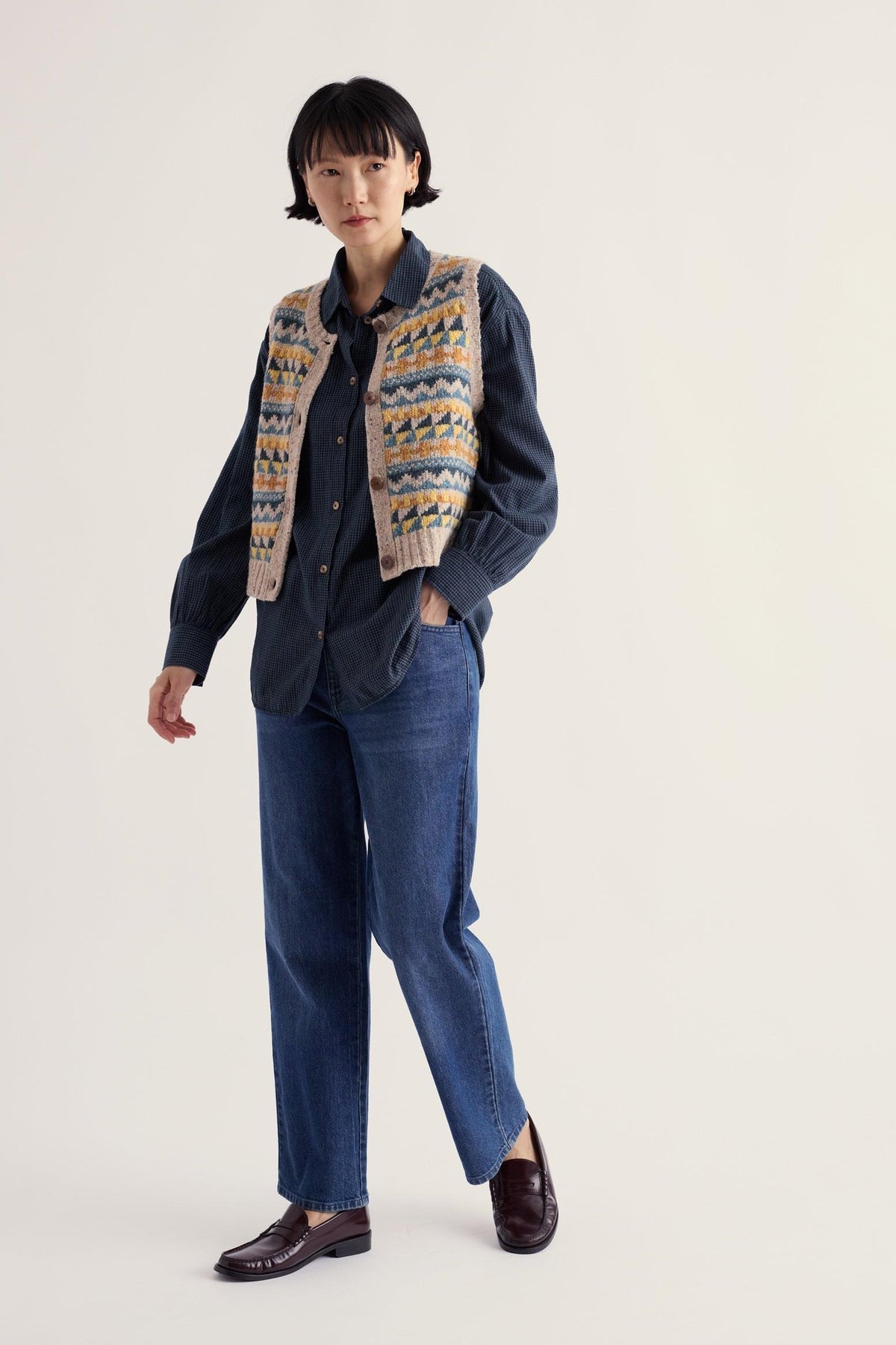 A woman modeling a Seasalt Cornwall patterned knit vest over a dark shirt and jeans against a plain background.