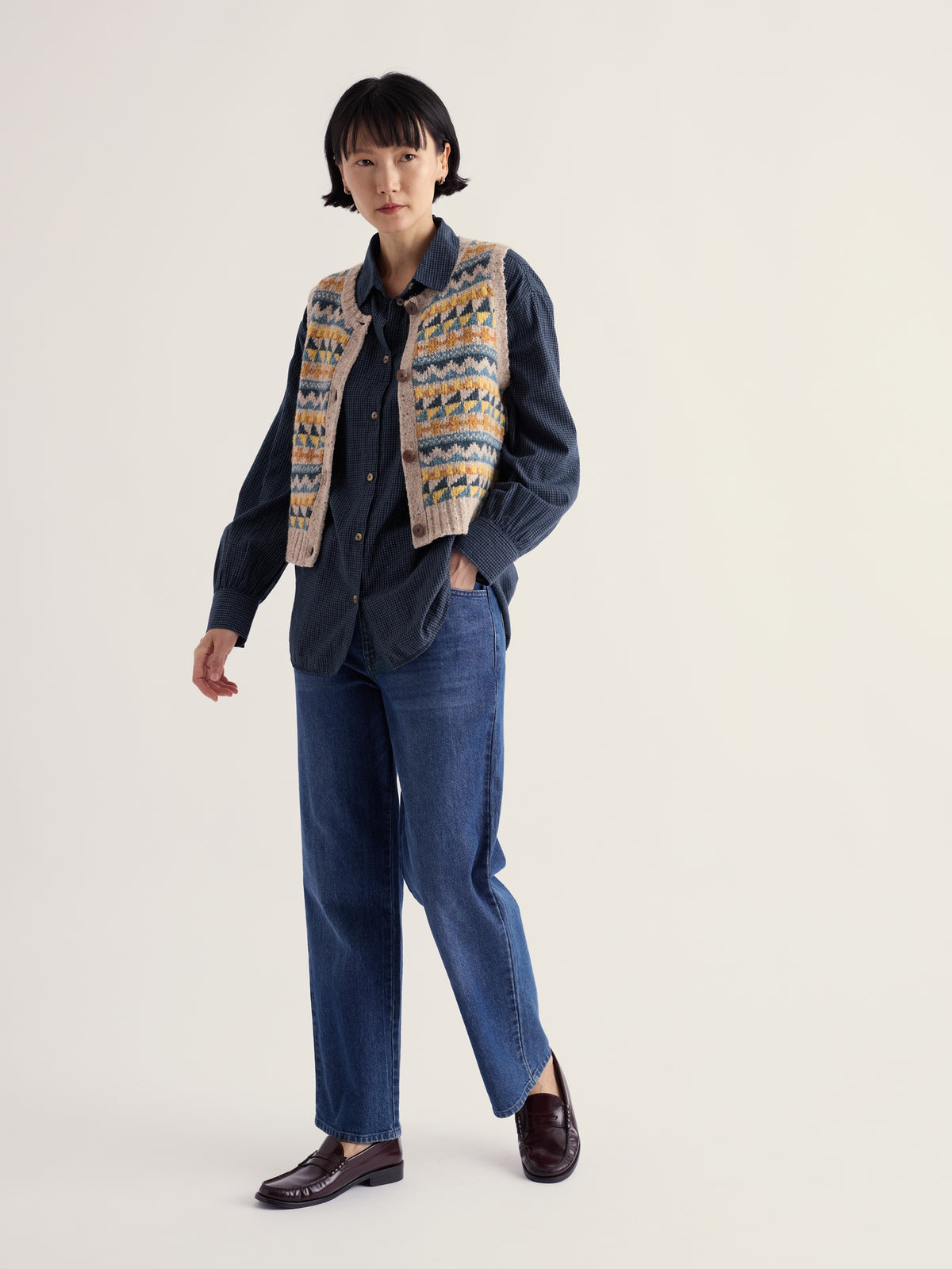 A woman modeling a Seasalt Cornwall patterned knit vest over a dark shirt and jeans against a plain background.