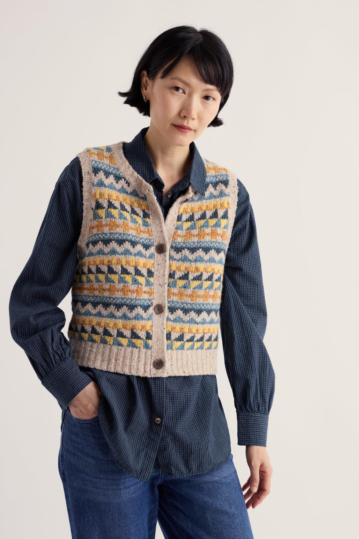 A woman wearing a beige Fairisle pattern knit vest over a dark blue plaid button-down shirt.