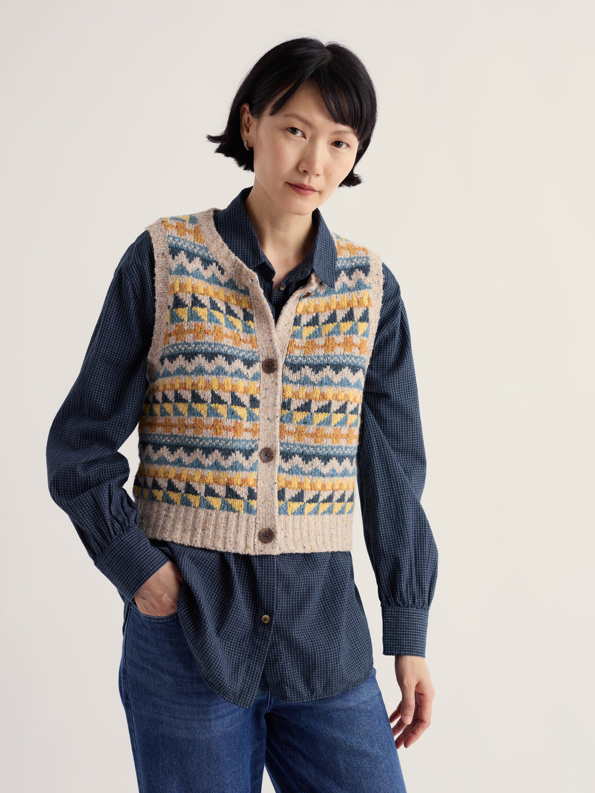 A woman wearing a beige Fairisle pattern knit vest over a dark blue plaid button-down shirt.
