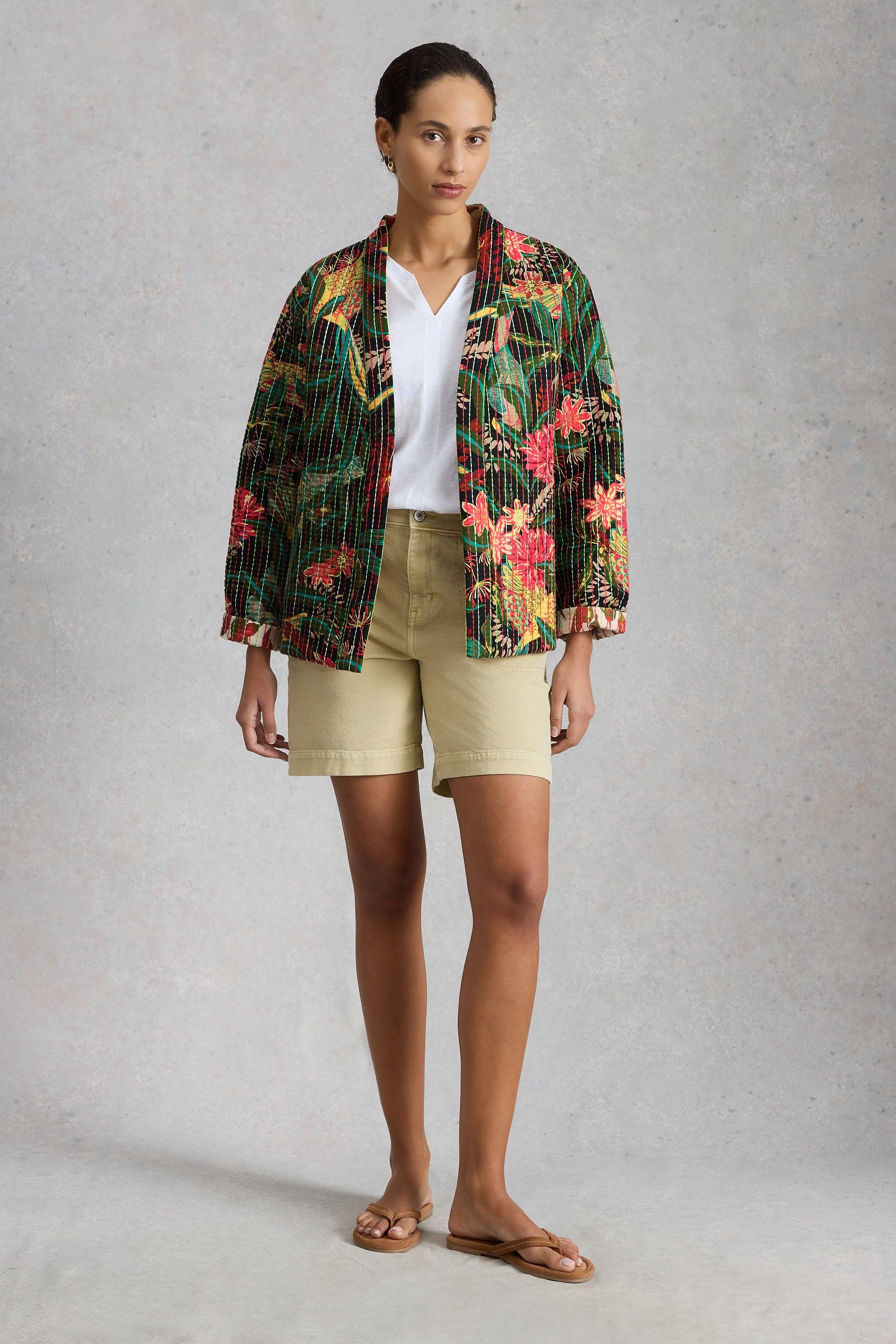 A woman posing in a floral patterned organic cotton jacket, white v-neck t-shirt, and khaki shorts.