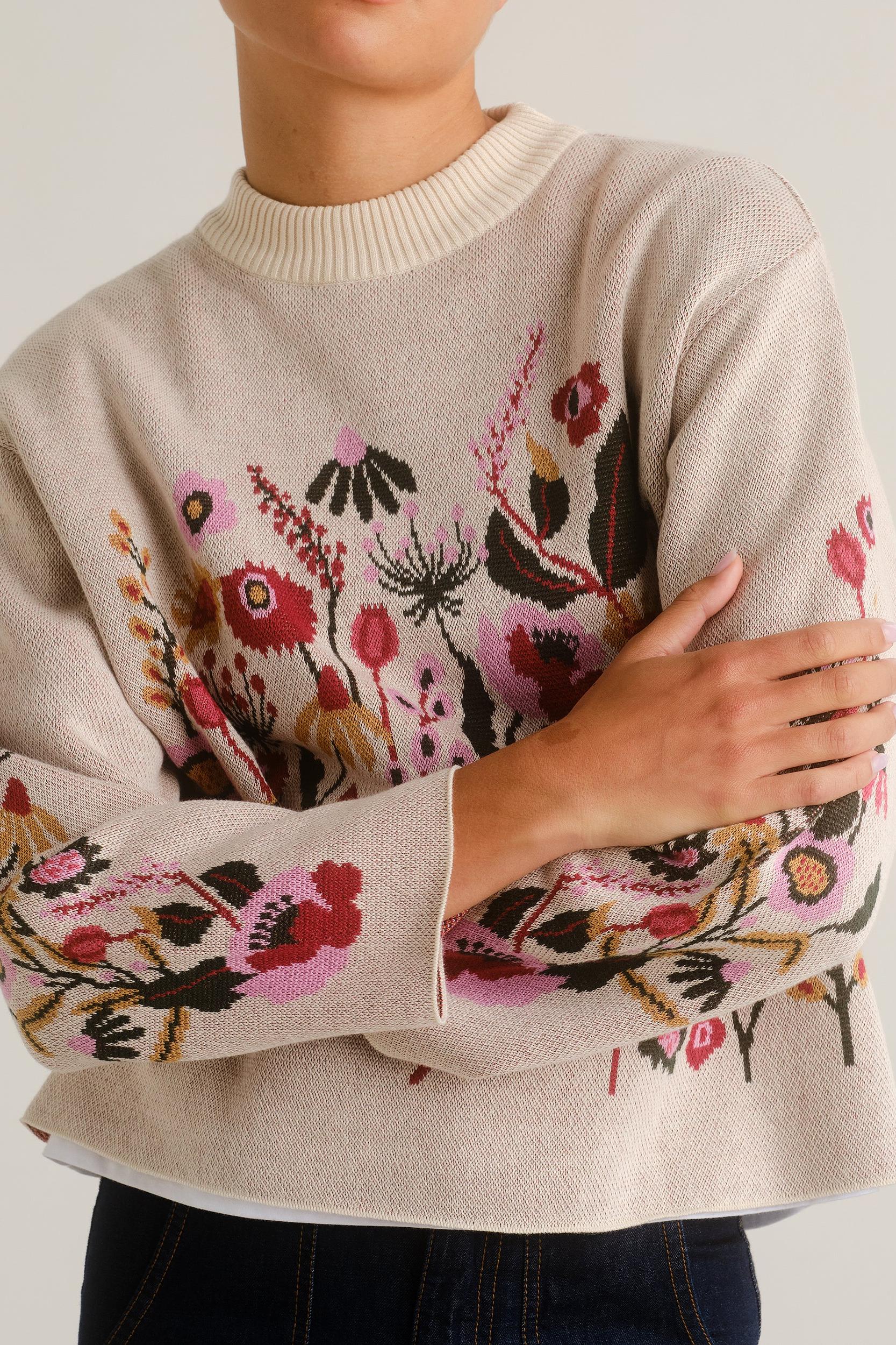 A close-up of a person wearing a cream-colored knit sweater with a colorful floral and botanical pattern in pink, red, green, and brown across the front and sleeves, paired with dark blue jeans.