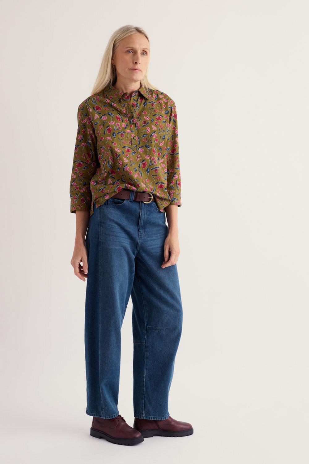 A woman wearing a long-sleeved floral organic cotton top tucked into loose blue jeans against a neutral background.