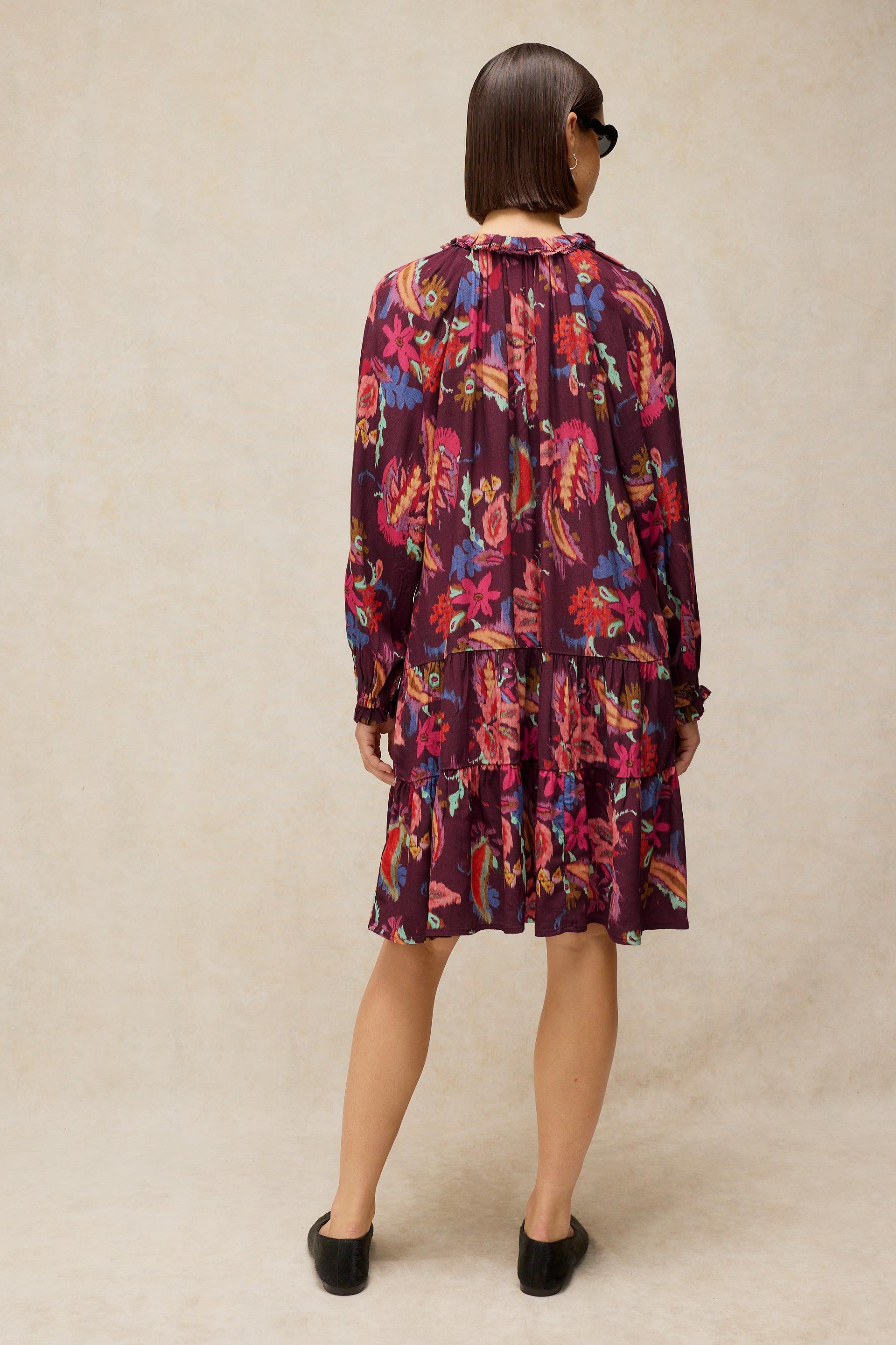 A woman viewed from behind wearing a long-sleeve tiered dress with a dense floral print in purple, pink, orange, and blue on a dark background, paired with black shoes against a textured beige backdrop.