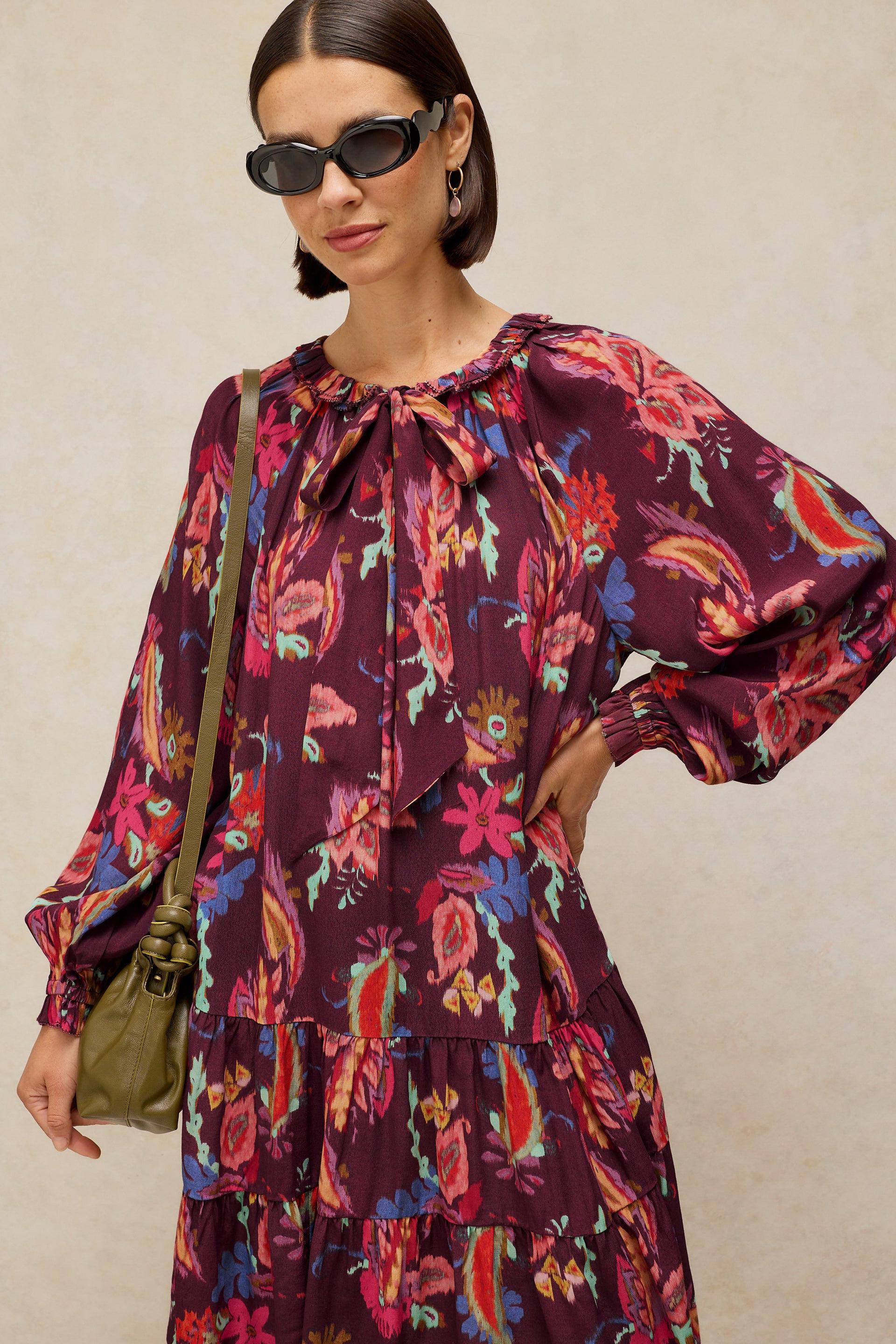 A woman modeling a long-sleeve tiered maxi dress with a vibrant paisley and floral print in deep purple, pink, red, and blue. She is wearing black sunglasses and carrying an olive green shoulder bag against a textured beige background.