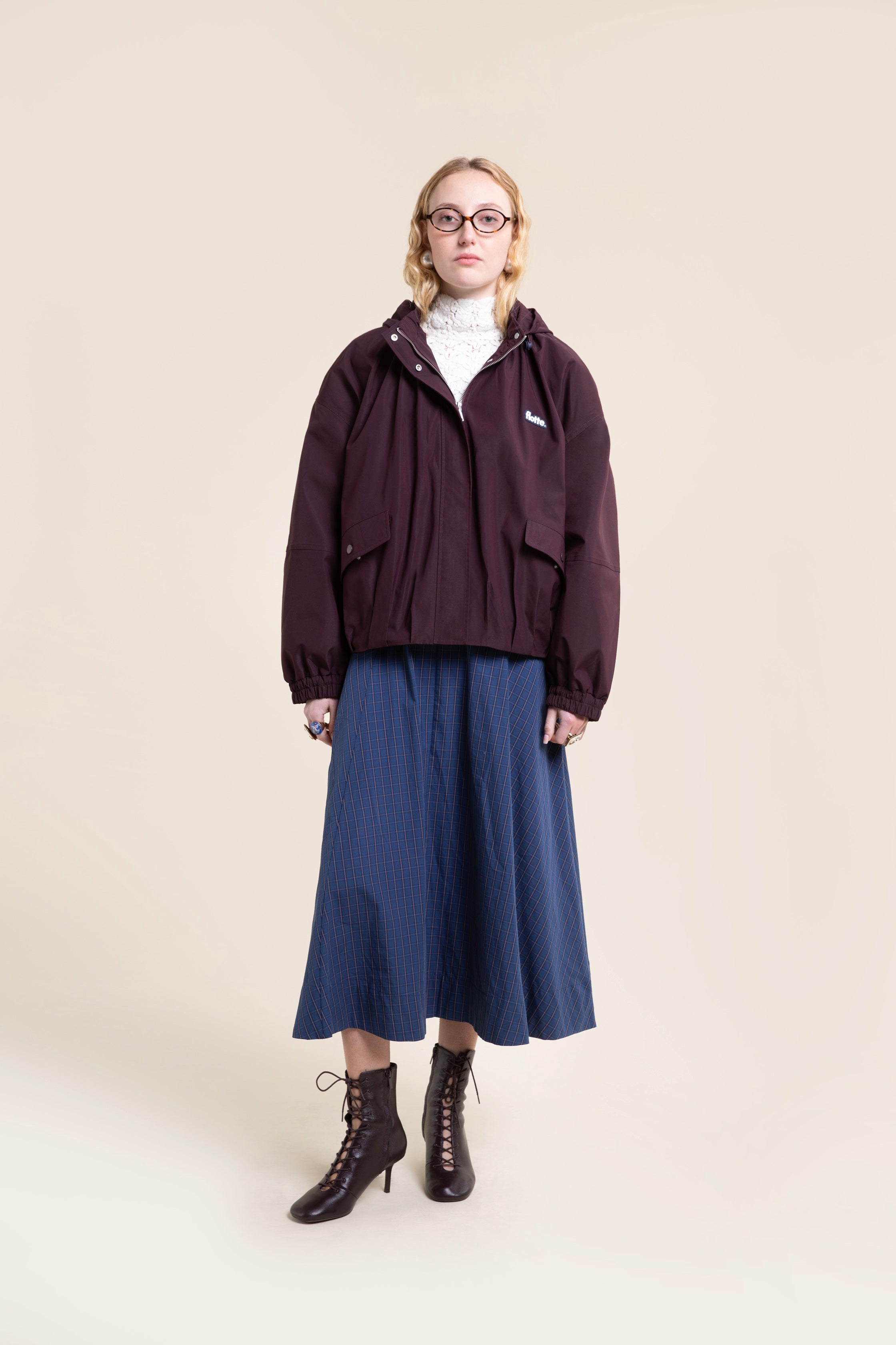 A woman modeling a stylish dark purple Flotte waterproof coat over a white top and blue plaid skirt.