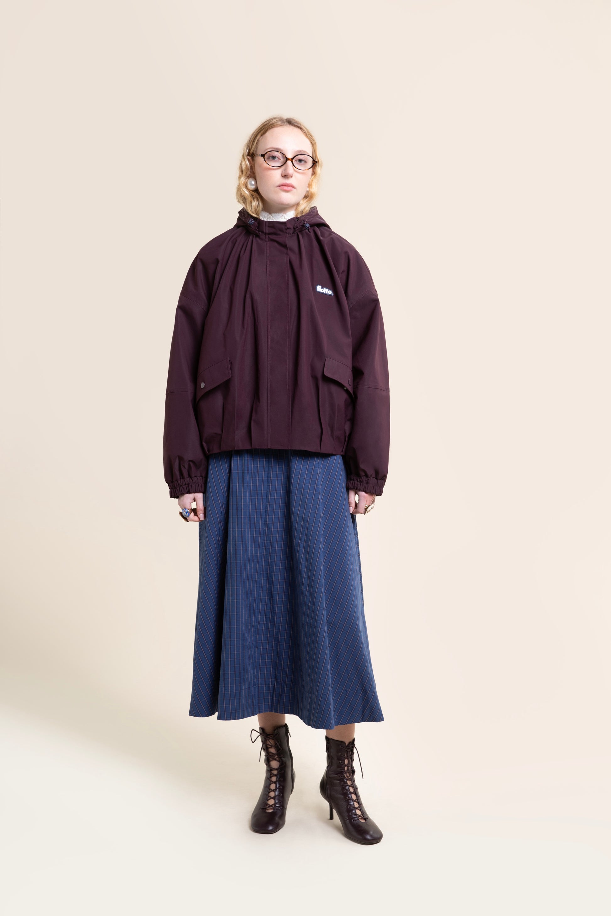 A woman wearing a dark purple Flotte waterproof coat over a blue plaid skirt against a beige studio background.