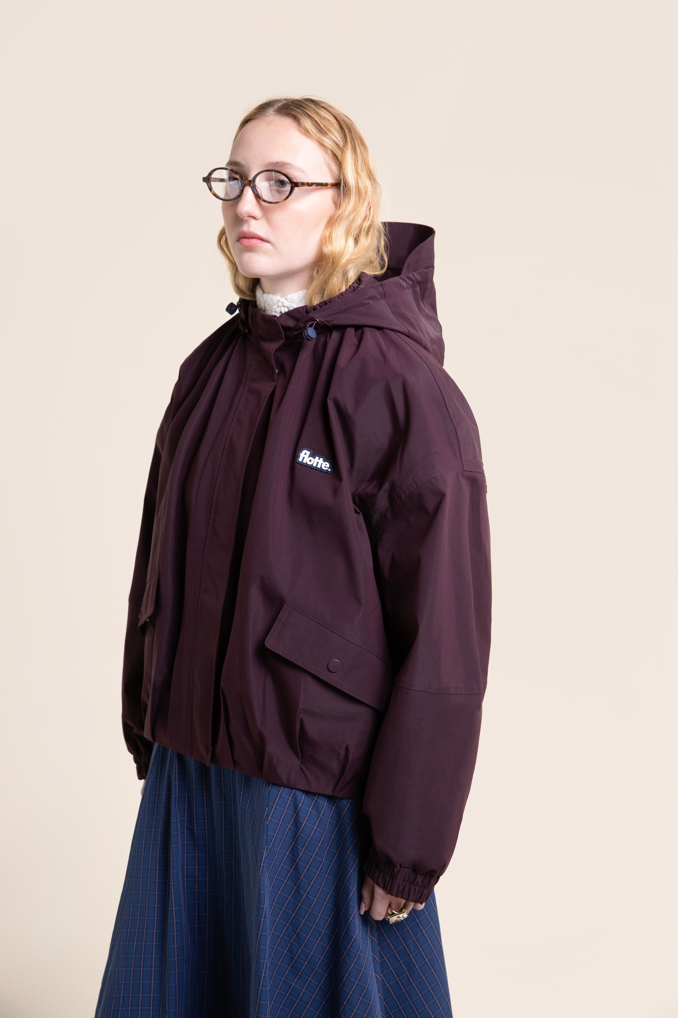 A woman wearing a dark purple Flotte waterproof coat over a blue plaid skirt against a neutral background.