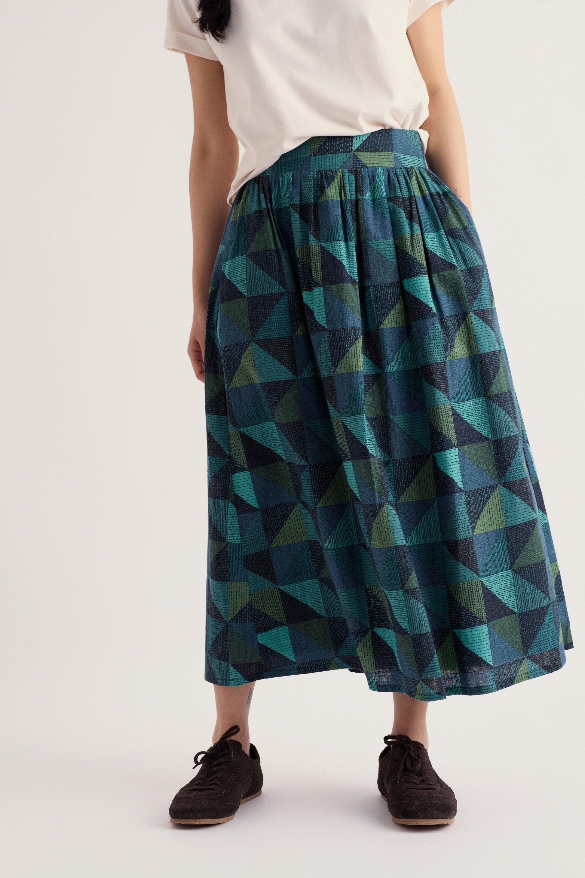 A person wearing a pleated midi skirt with a geometric triangle pattern in teal, green, and navy, paired with a white top and dark brown shoes, against a plain light gray studio background.