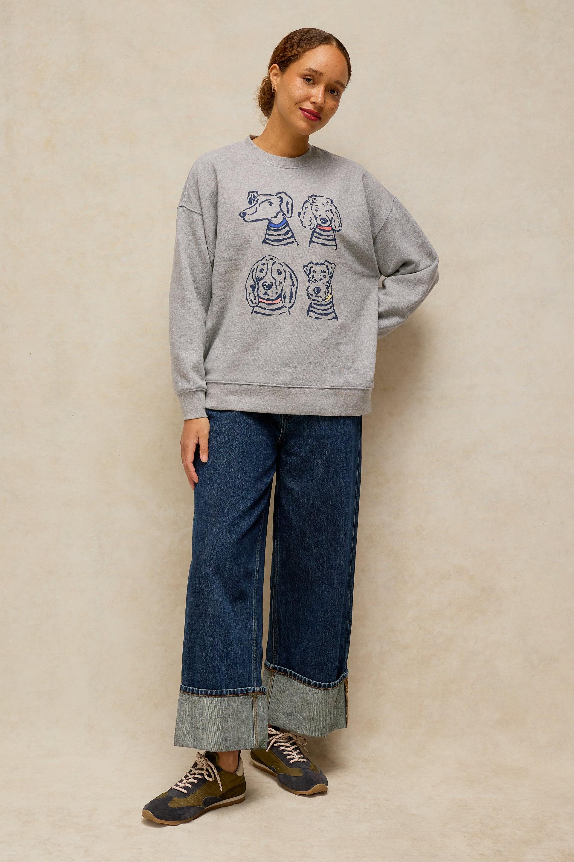 A woman modeling a light gray crewneck sweatshirt with a graphic print of four dogs, paired with wide-leg blue jeans and brown sneakers, standing against a textured beige wall.