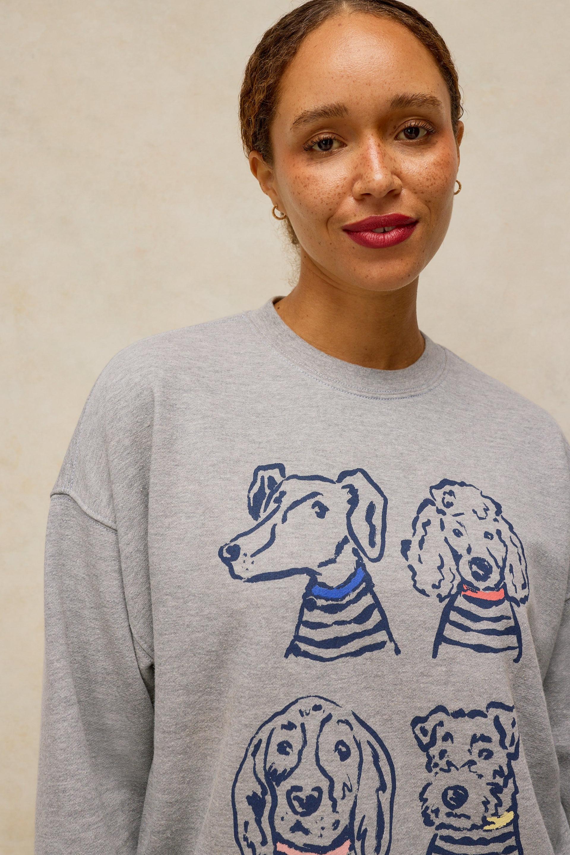 A woman wearing a light gray crewneck sweatshirt with a graphic print of four dogs wearing striped shirts in blue, navy, and red, set against a neutral beige background.
